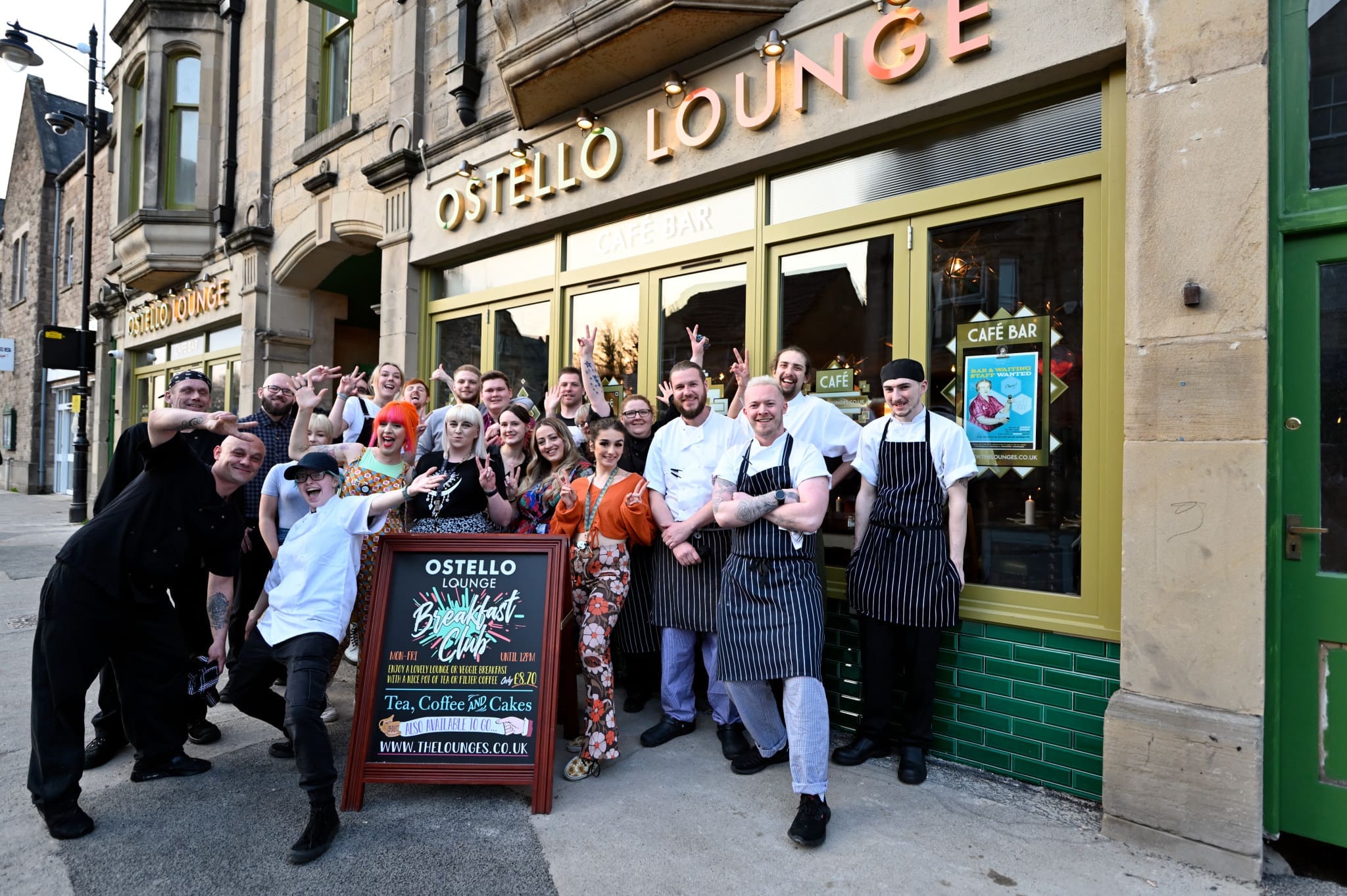 A large group of smiling people, including some in chef uniforms, pose together outside Ostello Lounge Café Bar, standing around a sign advertising Ostello. The setting is a city street lined with stone buildings.