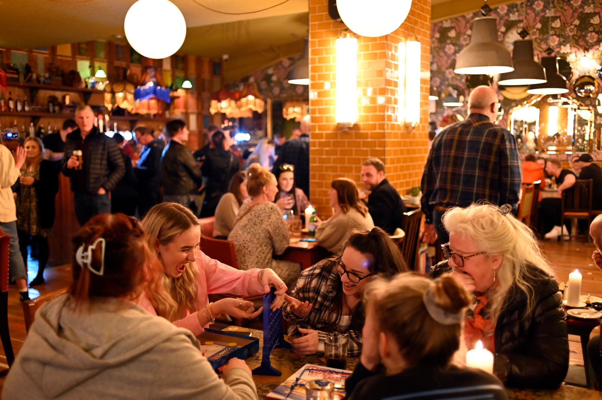 A lively, crowded Ostello bar with people socializing, drinking, and playing board games at tables. Warm lighting and colorful decor create a cozy atmosphere as four women in the foreground focus intently on a board game.