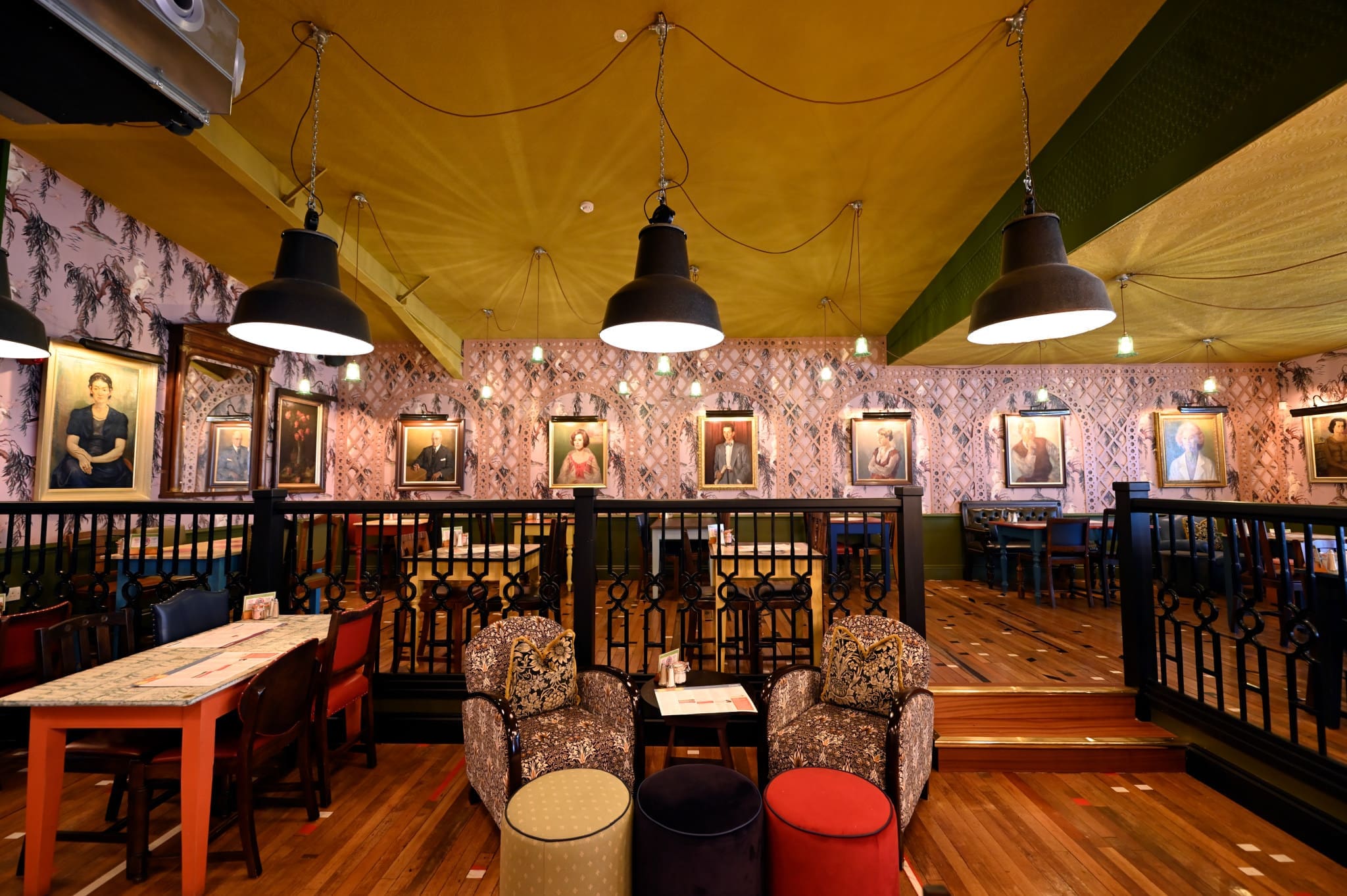 A cozy, eclectic café interior reminiscent of an Ostello, with patterned wallpaper, large hanging lights, framed portraits on the walls, mismatched chairs, and wooden floors creating a warm, inviting atmosphere.