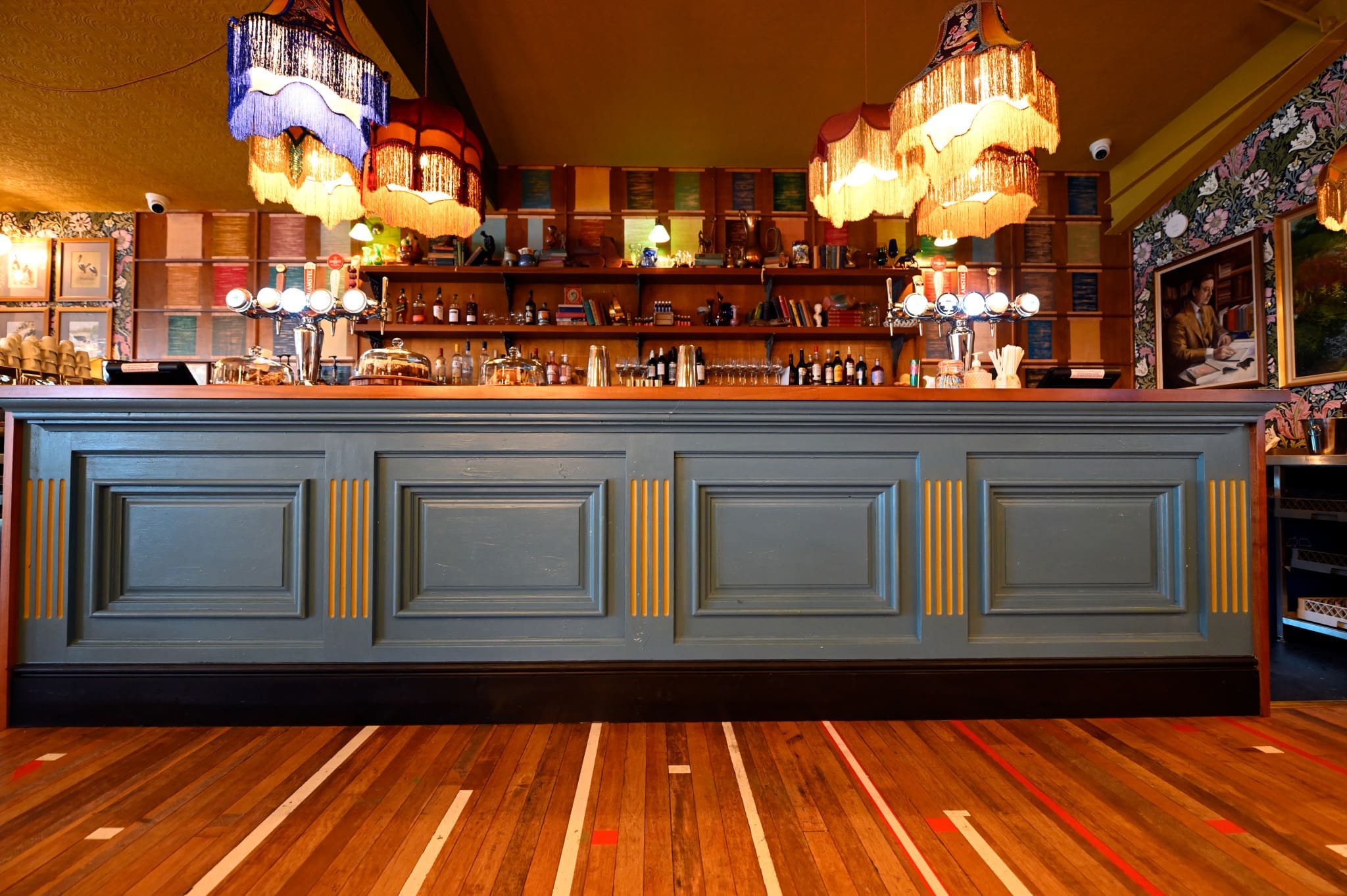 A stylish Ostello bar with a blue and gold front panel, wooden floor, hanging vintage lamps, and shelves of bottles and glasses behind the counter, decorated with wallpaper and framed art.