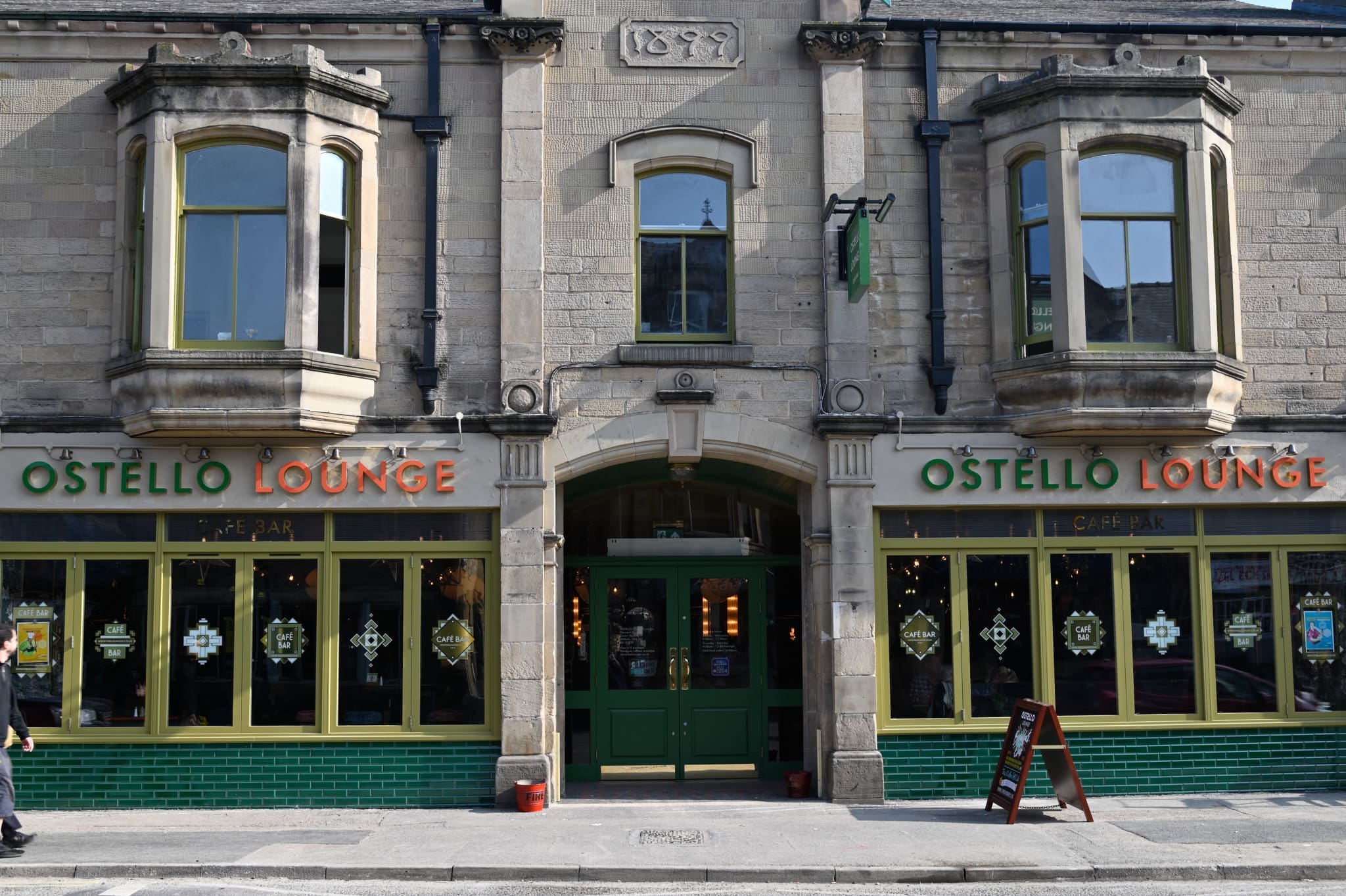 Front view of Ostello Lounge, a café-bar in a stone building with large bay windows, green doors, and signature orange-and-green Ostello signage. Decorative lights and seating are visible inside through the windows.