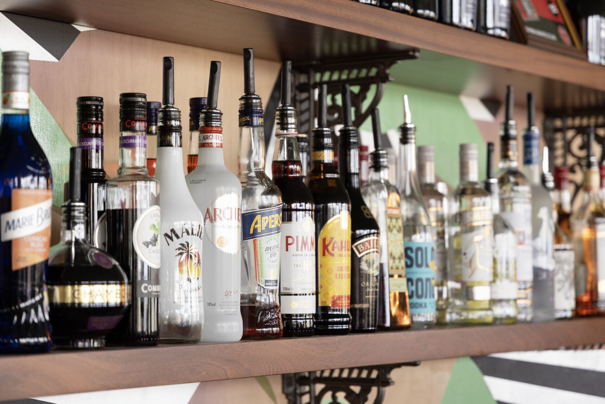 Bottles of various alcoholic beverages, including perico and other brands of vodka, rum, whiskey, and liqueurs, are lined up on a wooden shelf backed by a colorful mural with geometric patterns.