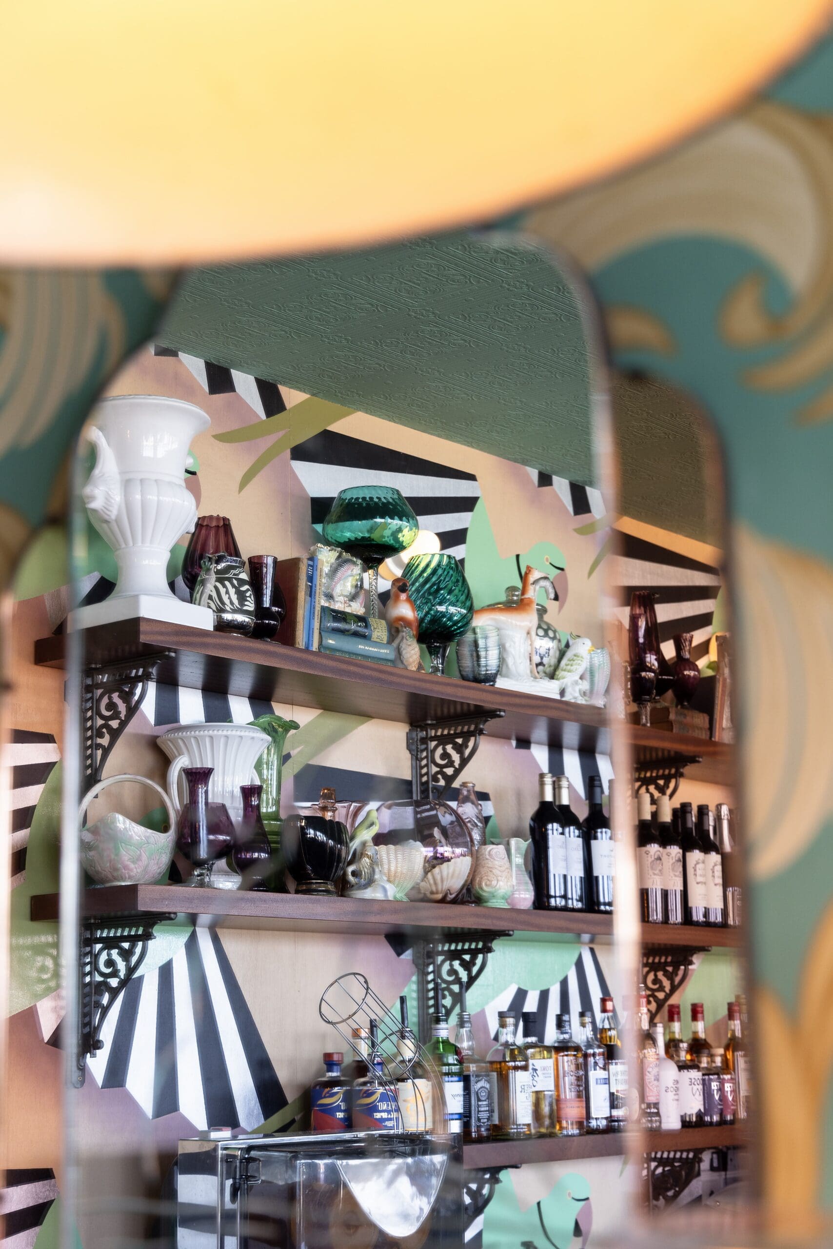 Shelves with colorful vases, ceramic figures, bottles, and bowls are reflected in a mirror. The background features a bold, perico-inspired geometric mural with green, black, white, and pink patterns.
