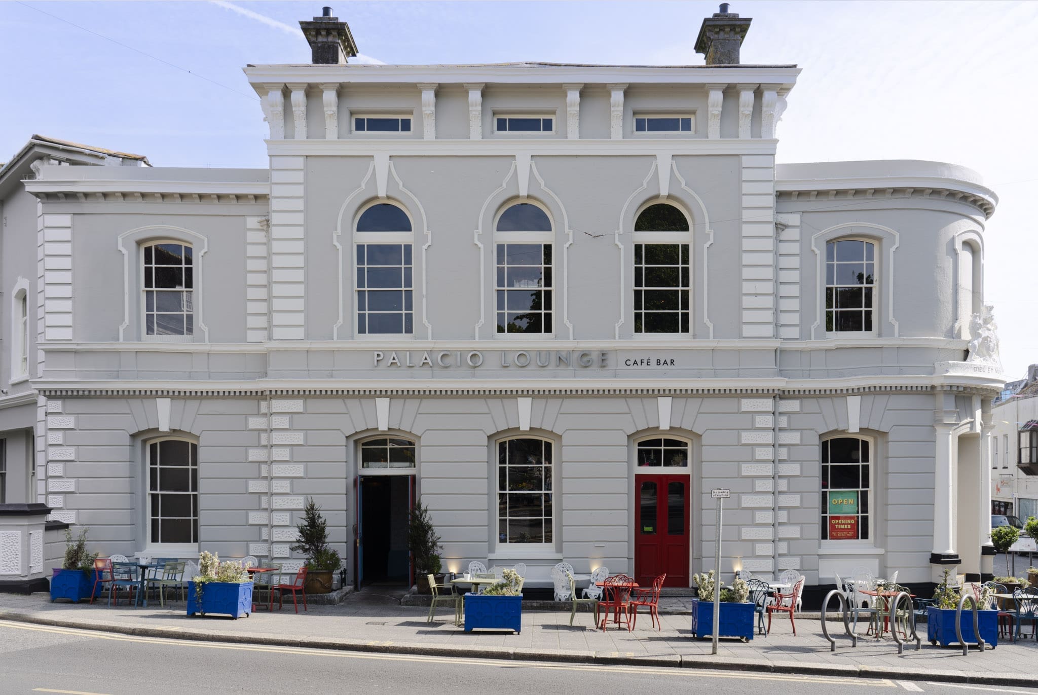 A two-story white Palacio building with arched windows and a red door, featuring a sign that reads "Palacio Lounge Café Bar." Colorful outdoor tables and chairs line the sidewalk in front.