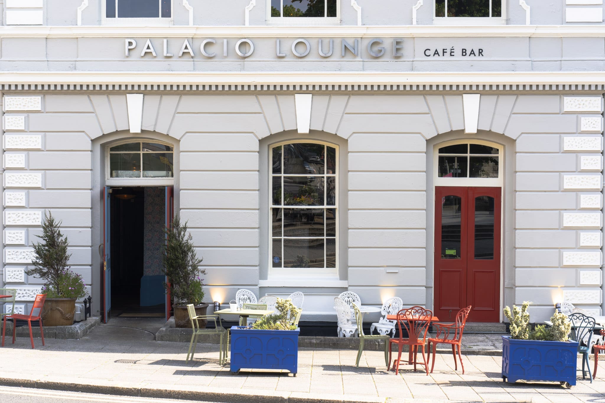 The exterior of Palacio Lounge café bar features signature Palacio charm—two red doors, arched windows, and outdoor seating with colorful chairs, tables, and vibrant potted plants on a sunny day.