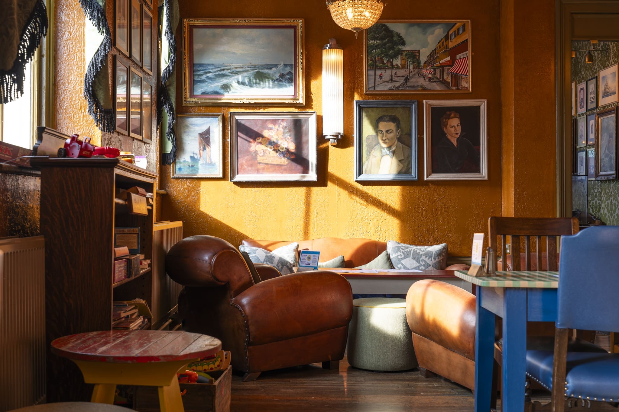 Cozy café interior with a brown leather armchair, small round table, and bookshelf evokes the charm of a mini Palacio. Sunlight streams in onto framed paintings and portraits, creating a warm, inviting atmosphere.