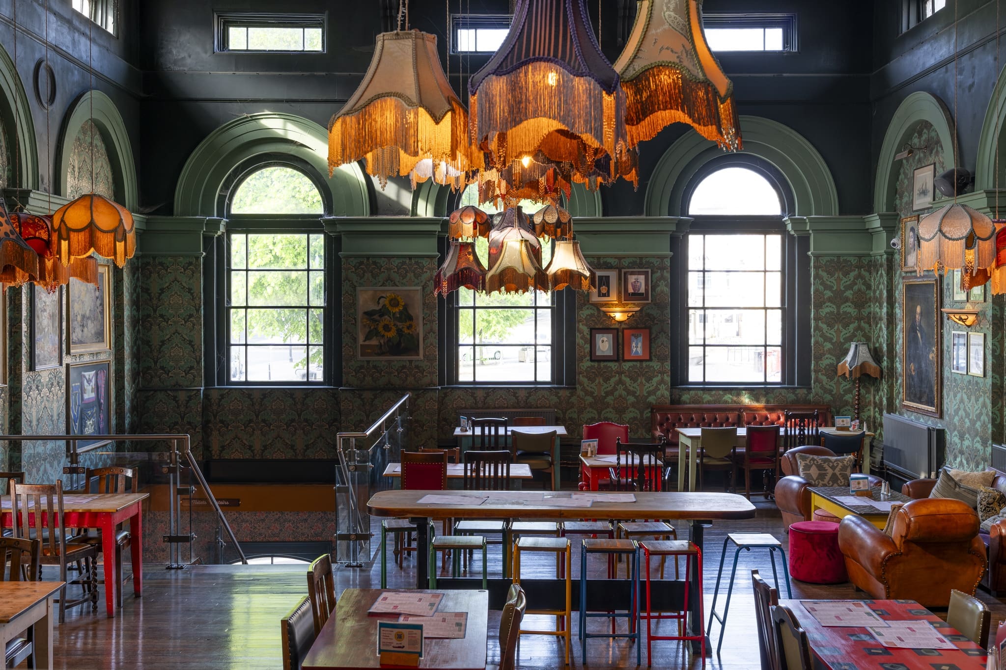 A cozy, vintage-style café with eclectic lampshades hanging from the ceiling, patterned wallpaper, and large arched windows reminiscent of a palacio. Mismatched chairs and tables pair with framed art while natural light fills the room.