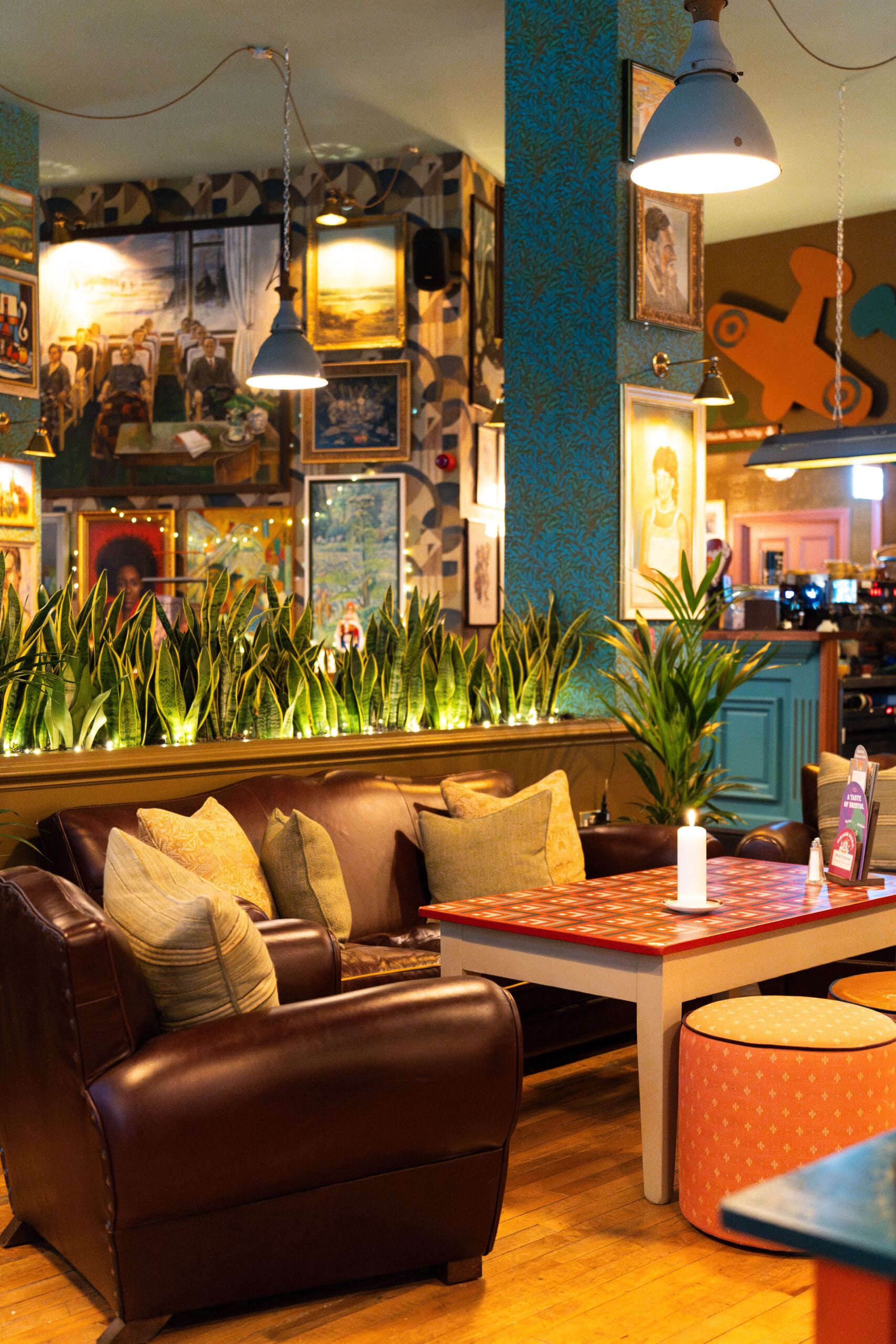 A cozy café interior featuring a brown leather sofa, colorful cushions, a candlelit table, green plants, framed artwork—including a Palomo print—on the walls, and warm lighting creating a welcoming atmosphere.