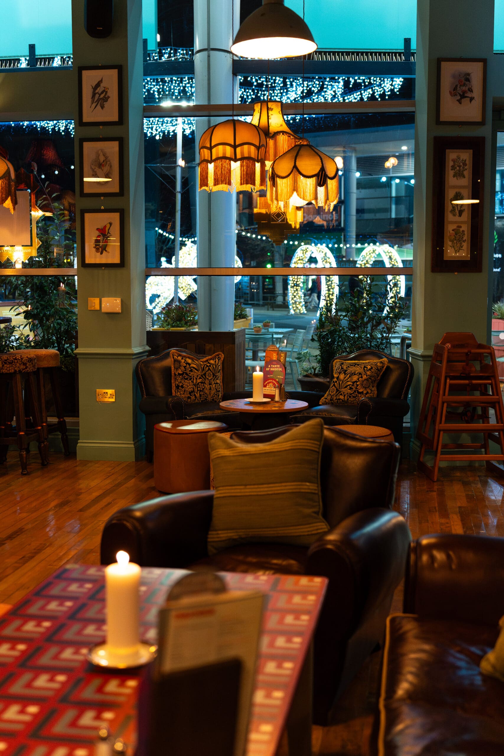 Cozy cafe interior with leather chairs and lounge areas, a candle on a table, vintage lamps, framed art on walls, and large windows showing festive lights outside. Warm, inviting atmosphere with soft lighting.