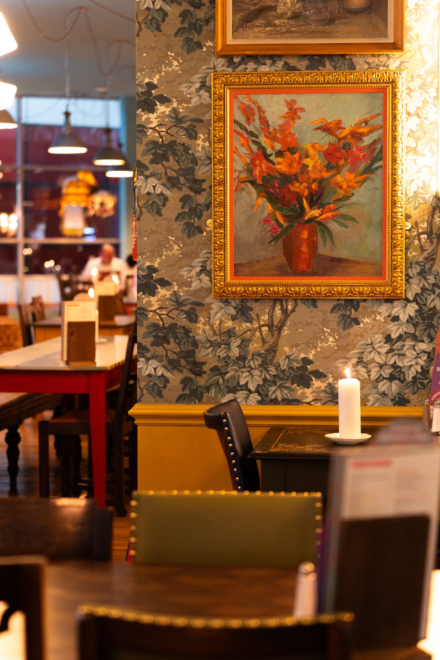 A warmly lit café interior with floral wallpaper, a framed painting of a vase with flowers, wooden tables and chairs, a lit candle on a table, palomo touches in the decor, and hanging lights in the background.