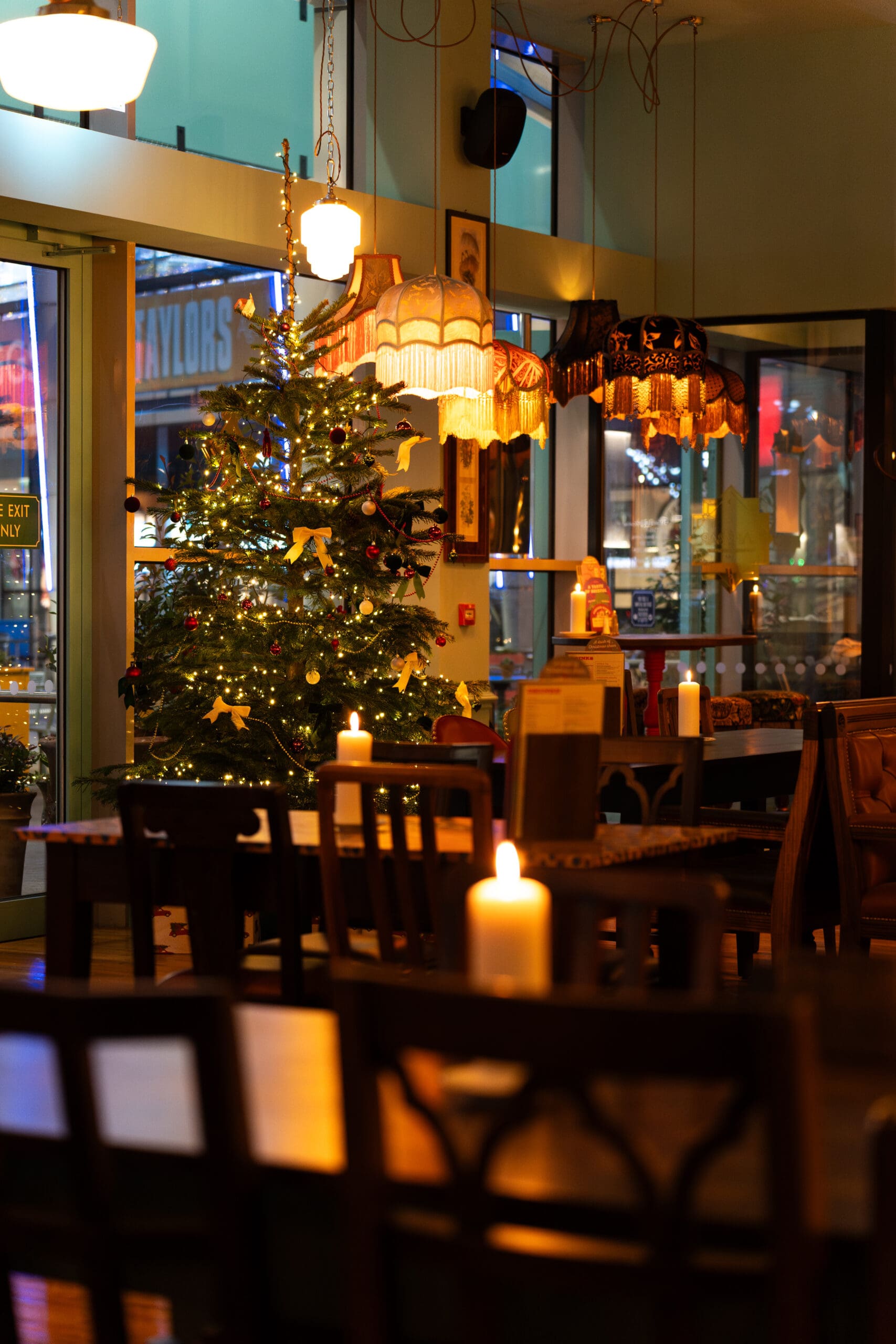 A warmly lit lounge café decorated for Christmas, featuring a lit tree with ornaments and lights, several glowing candles on tables, and vintage hanging lamps. The atmosphere is cozy and inviting.