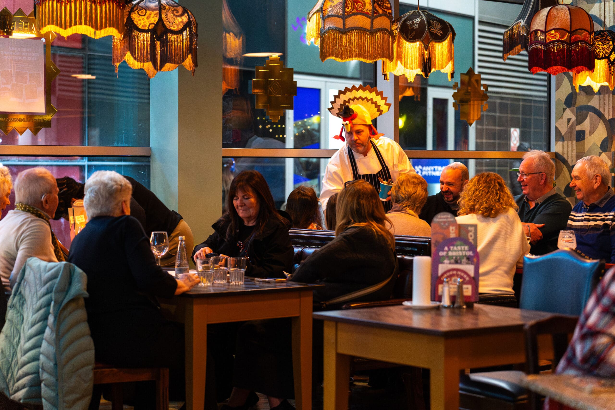 A group of people sit around tables in a cozy restaurant. A smiling chef wearing a feathered palomo headdress talks to diners. Warm lighting from decorative lamps creates a welcoming atmosphere.