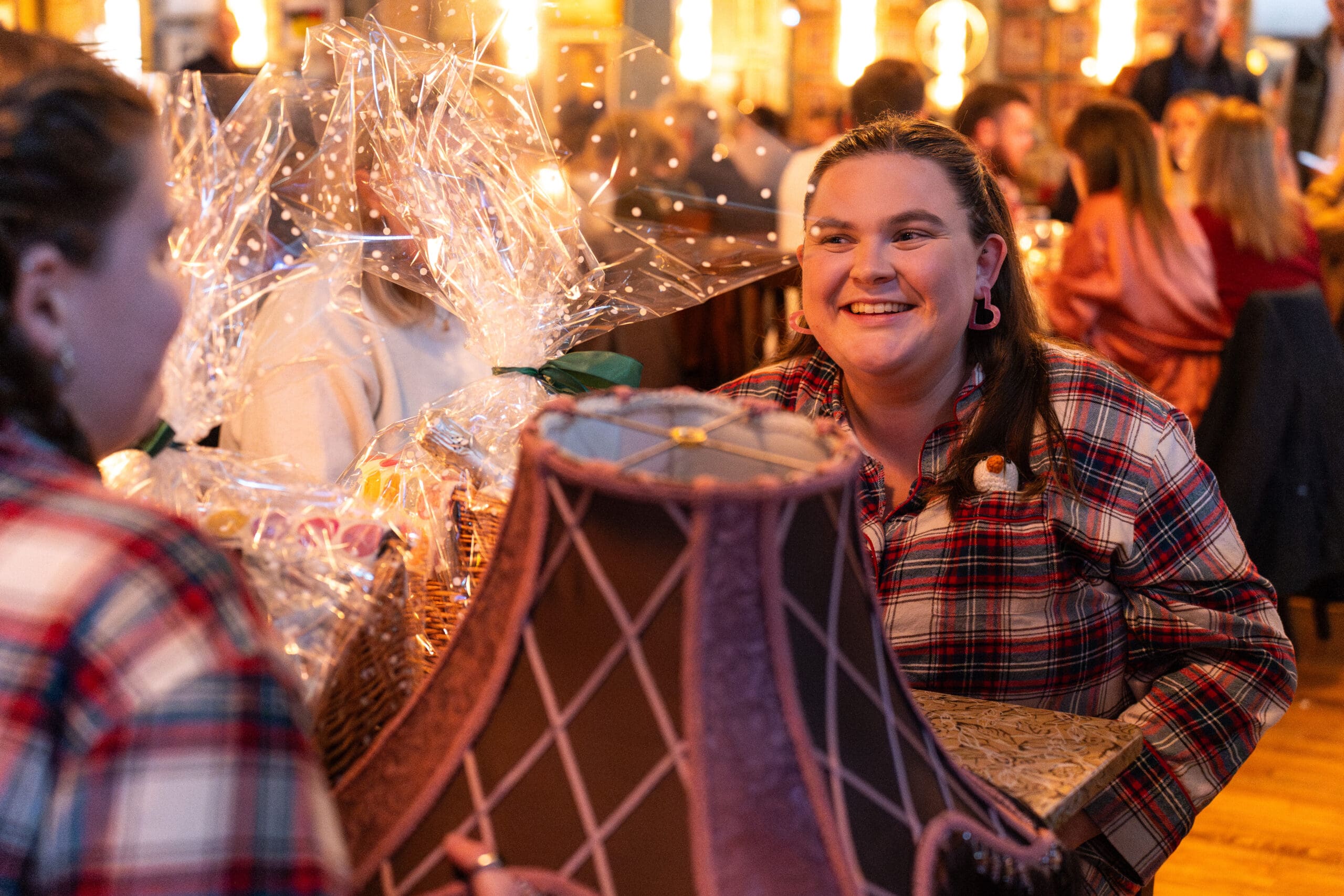 A smiling woman in a plaid shirt sits at a table in a warmly lit Palomo restaurant, holding a vintage-style lampshade and surrounded by wrapped gift baskets. Other people chat in the background.