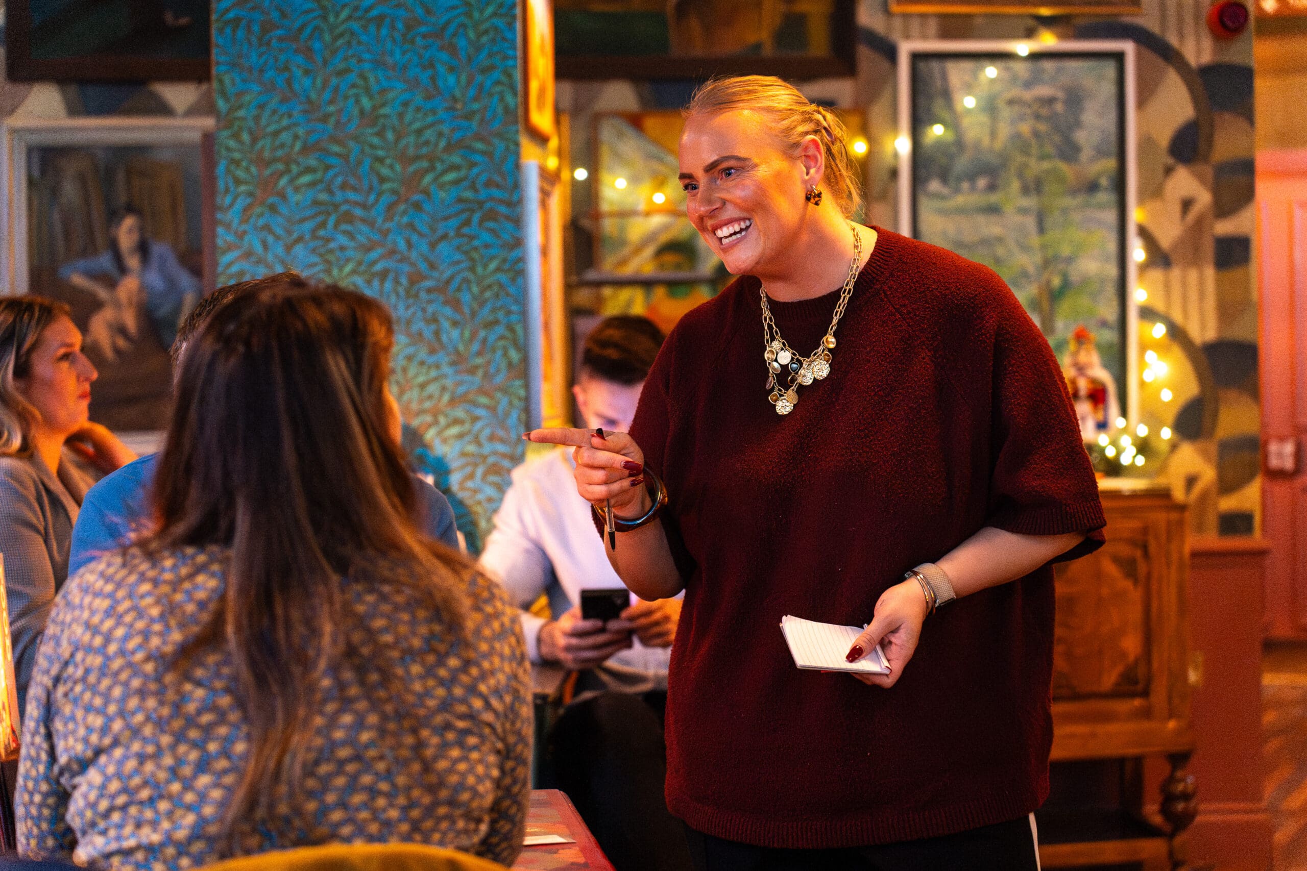 A smiling waitress holding a notepad talks to a customer at Palomo, a cozy, warmly lit restaurant. Other diners sit at tables, and framed art and patterned wallpaper decorate the background.