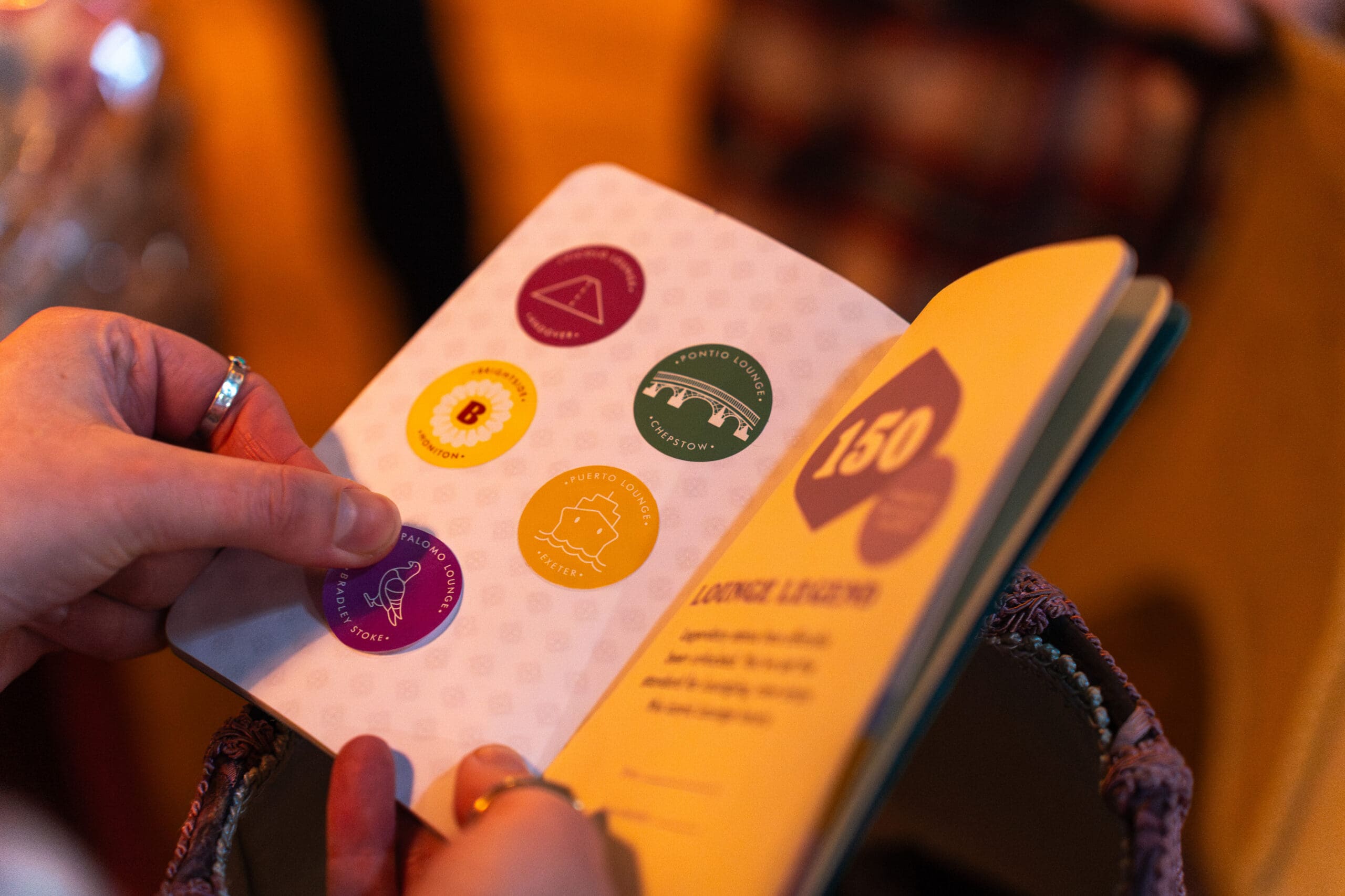 A person holds open a Palomo booklet displaying colorful circular stickers on one page and text with a yellow "150" graphic on the other, with a soft, warm background.