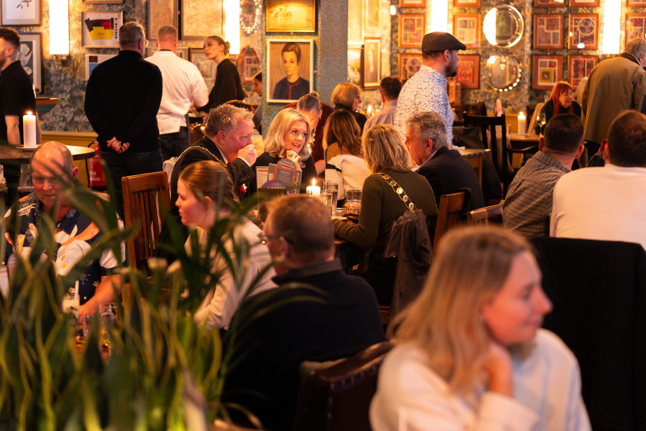 A lively palomo restaurant scene with people of various ages sitting at wooden tables, eating, drinking, and chatting. The warmly lit space features framed photos and mirrors on patterned walls, with plants in the foreground.