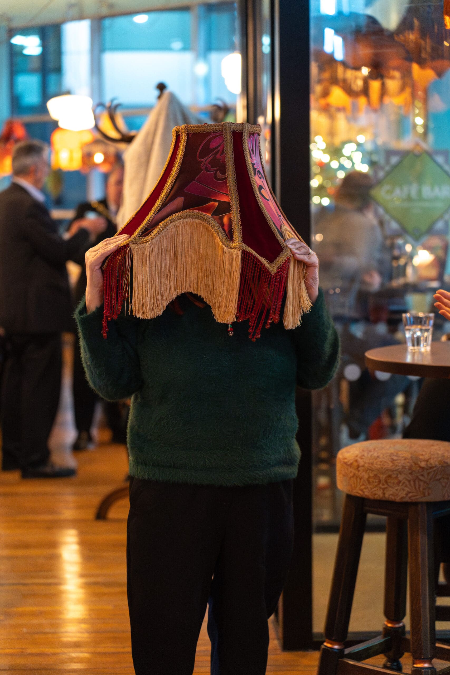 A person wearing a green Palomo sweater stands indoors with a decorative lampshade covering their head. People talk in the background of the lively, well-lit café or bar.