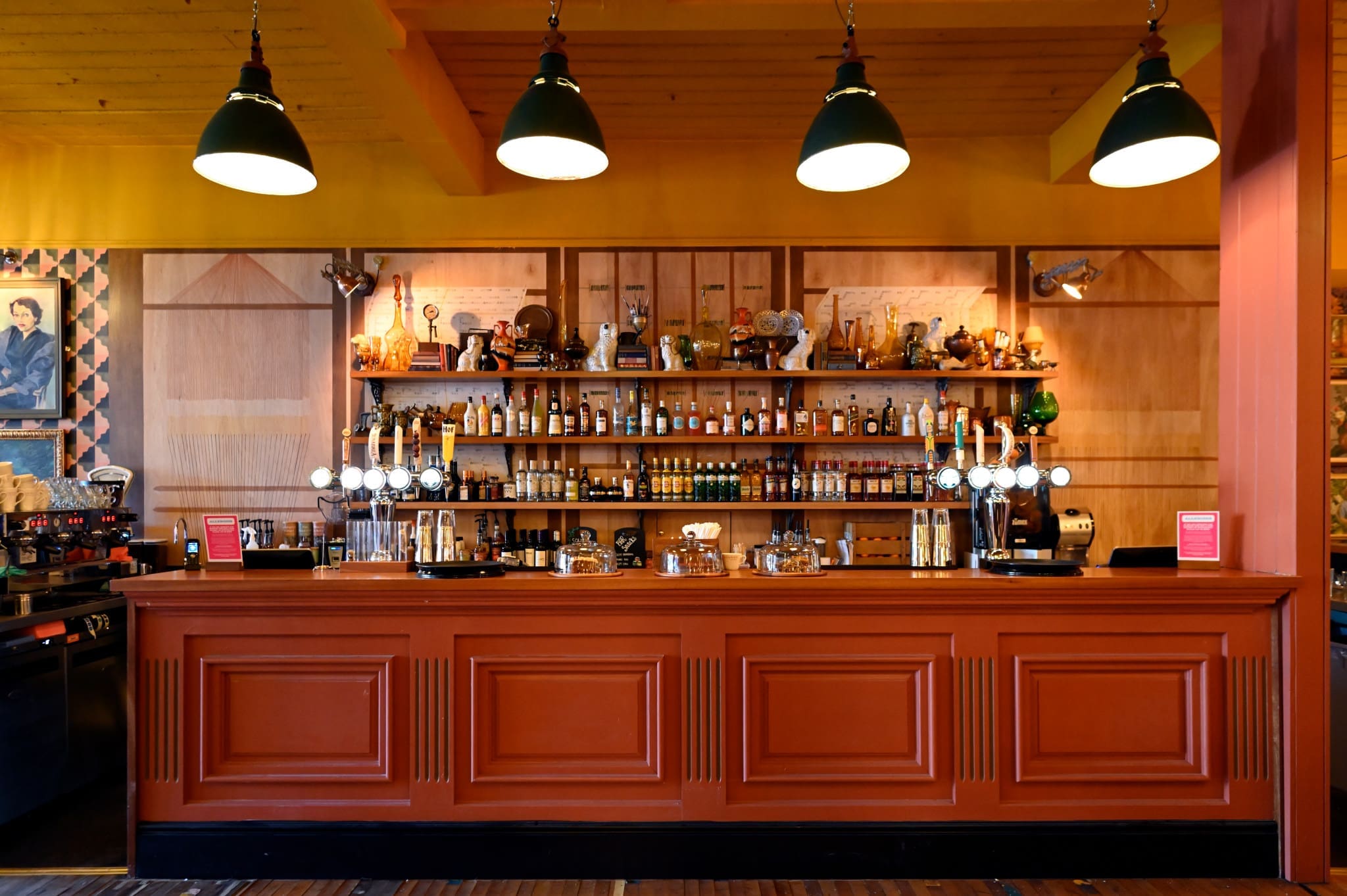 A brightly lit bar with an orange wooden counter, shelves filled with bottles, glasses, and barware, and three large green pendant lights above. The décor is warm, featuring various framed art and a nod to panadero craftsmanship in its woodwork.