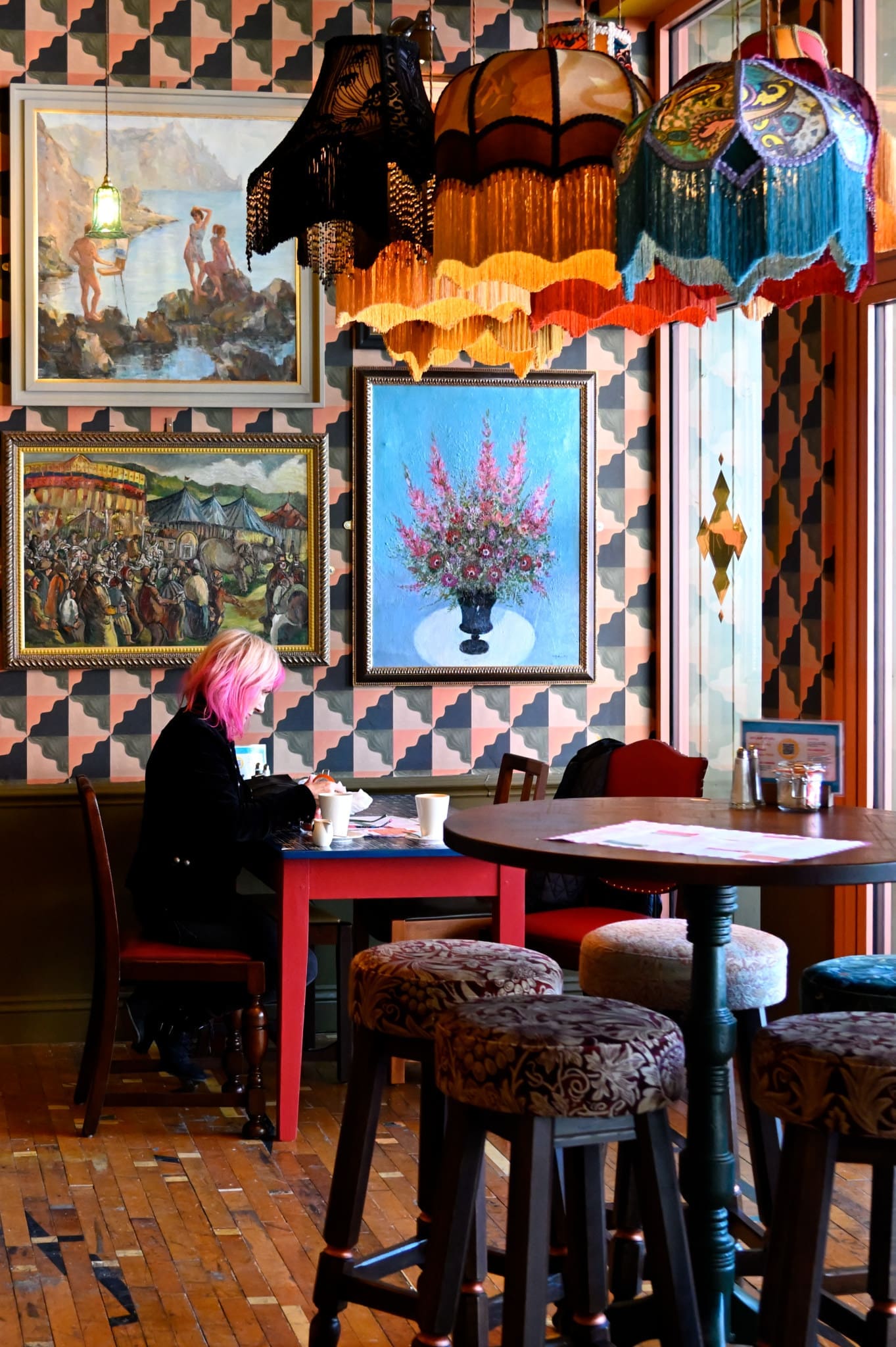 A panadero with pink hair sits alone at a red table in a colorful, eclectic café adorned with patterned wallpaper, vintage paintings, and ornate hanging lamps. Empty stools and a wooden floor fill the cozy space.
