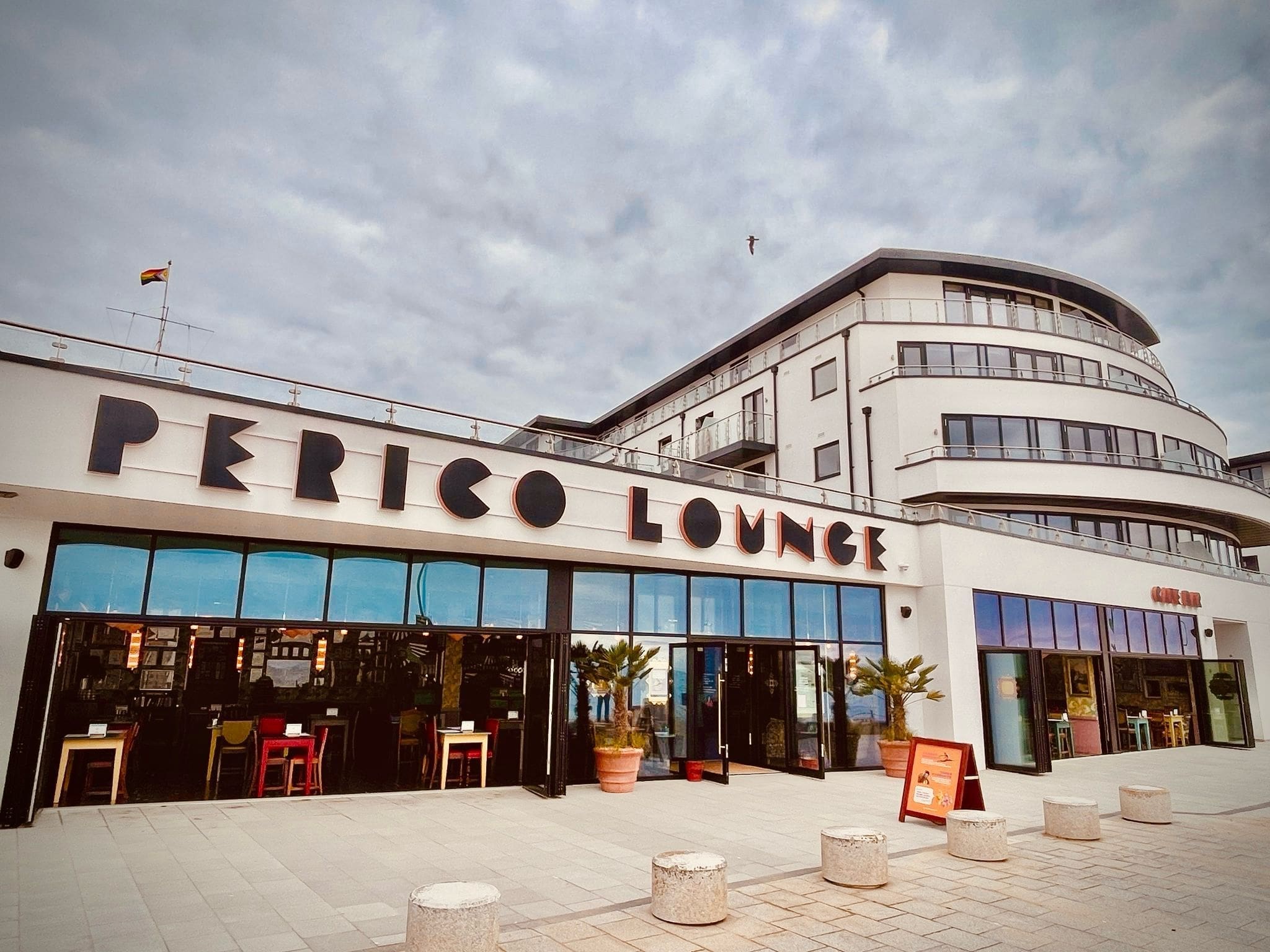 A modern building with large windows houses Perico Lounge. The bold "Perico" sign stands above an open, inviting entrance featuring outdoor seating, lush potted plants, and cloudy skies overhead.