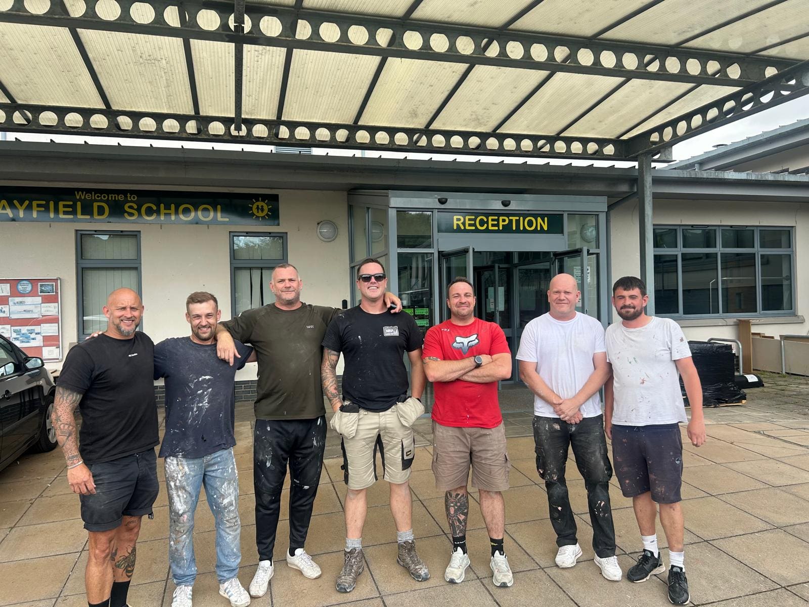 Seven men stand smiling in front of the entrance to a school building under a metal canopy, with a sign above reading "RECEPTION" and another sign saying "MAYFIELD SCHOOL" visible in the background.