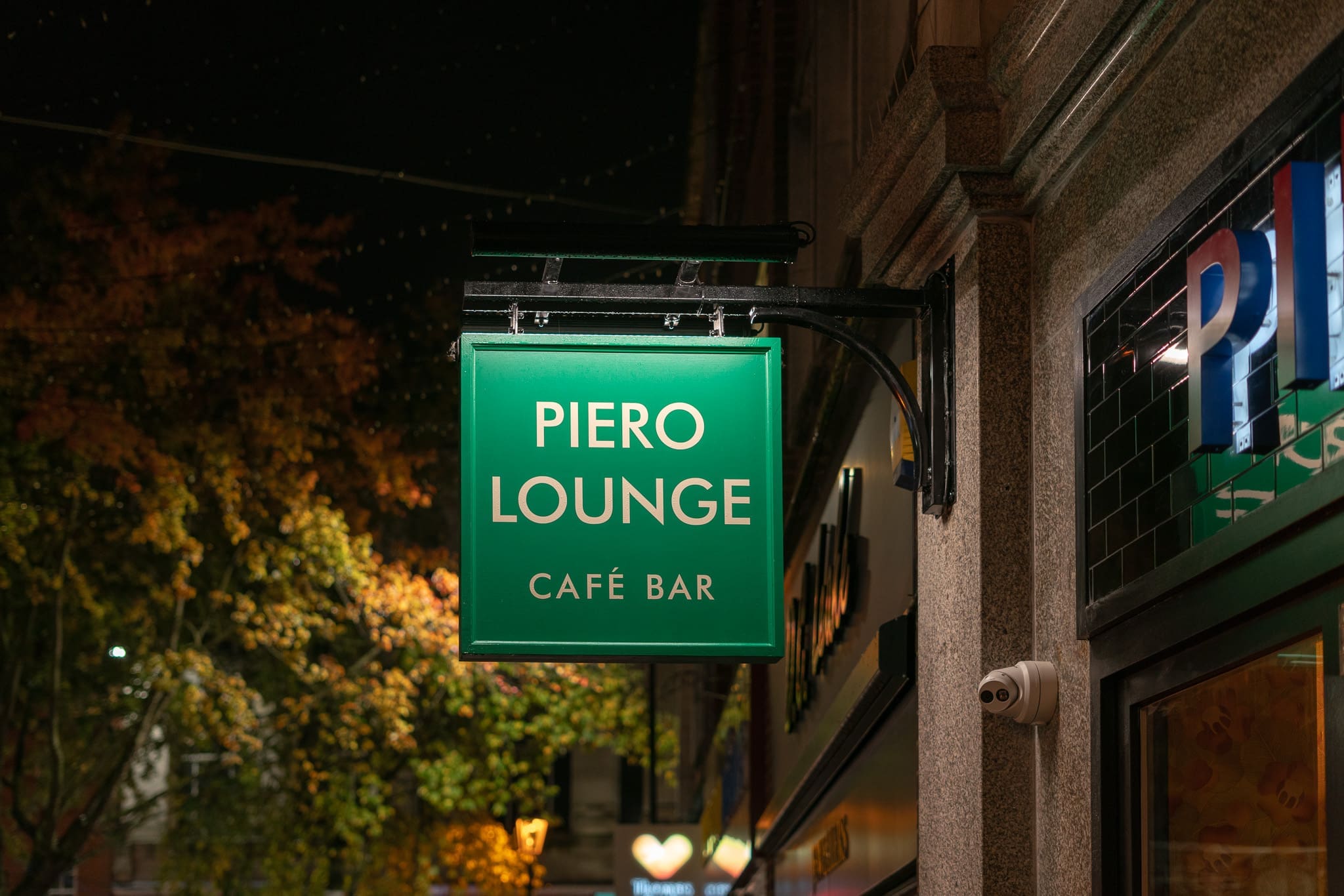 A green illuminated sign reading "PIERO LOUNGE CAFÉ BAR" hangs outside a building at night, showcasing the vibrant Piero atmosphere amid trees and soft streetlights in the background.