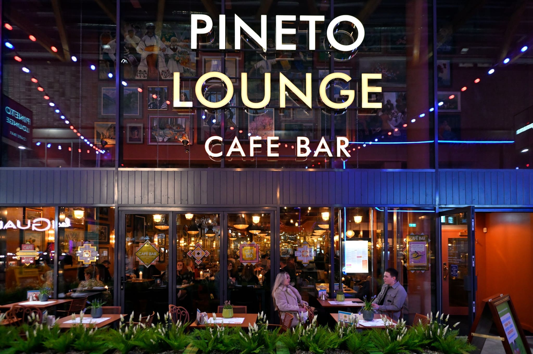 The exterior of Pineto Lounge Café Bar at night features large glass windows revealing people dining inside, while two people sit at a table outside, surrounded by green plants and colorful string lights.