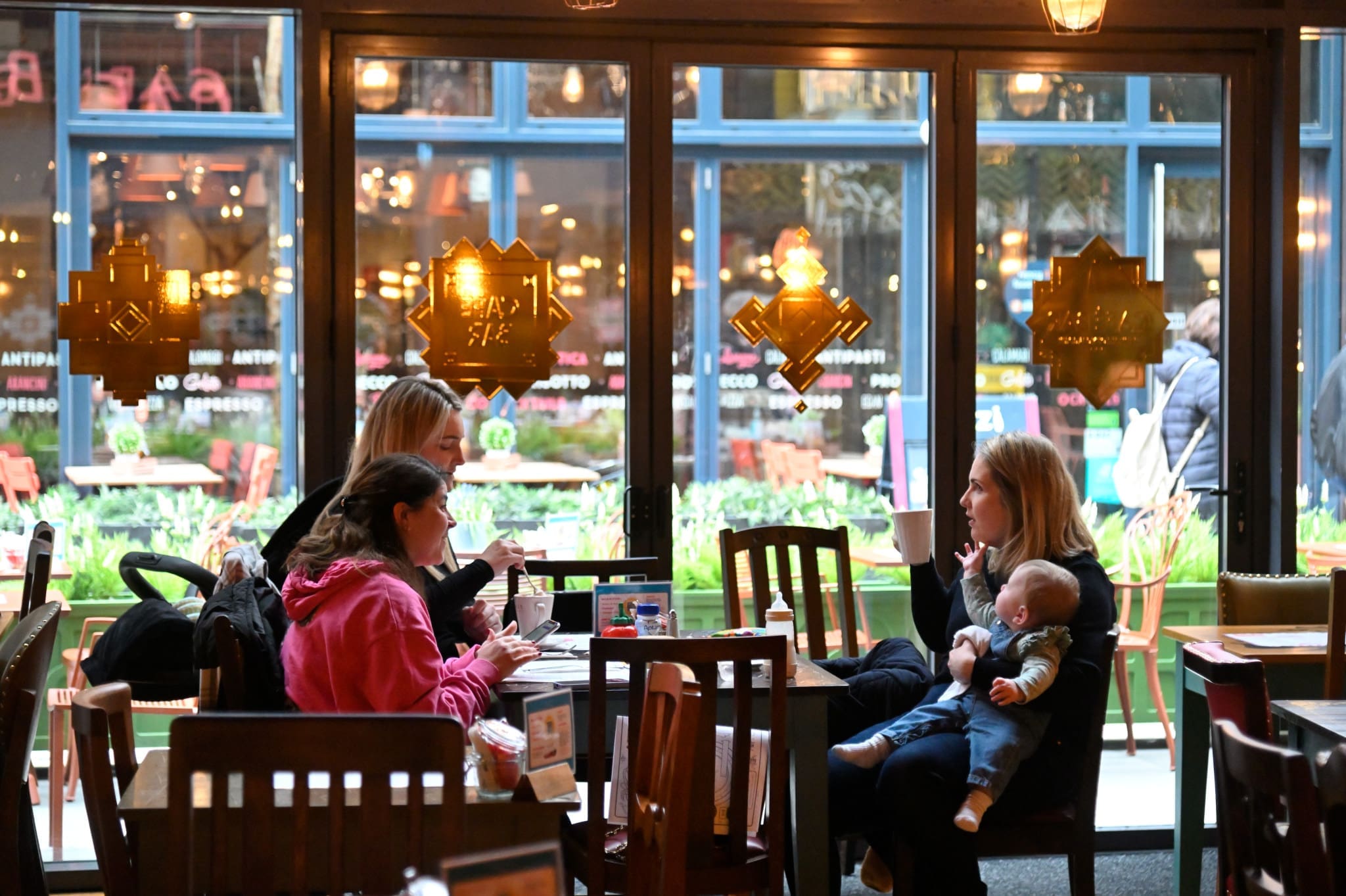 Three women sit at a café table by a large window in Pineto; one woman is holding a baby. Outdoor seating and pedestrians are visible through the glass. The café interior looks warm and inviting with wooden furniture and decorative glass decals.