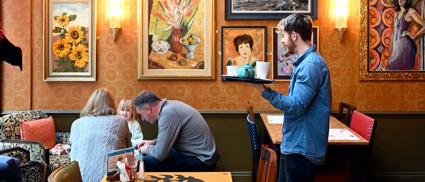 A waiter carries a tray with a teapot and cups in a cozy café in Pineto, decorated with colorful paintings, while a family with a child sits together at a table in the background.