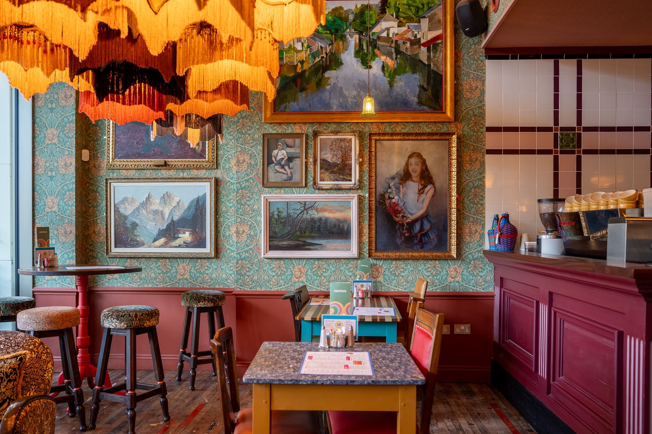 A cozy, eclectic café with vintage decor, colorful Pinto chairs, ornate wallpaper, framed paintings on the wall, warm lighting from a large fringed chandelier, and a counter with coffee machines on the right.