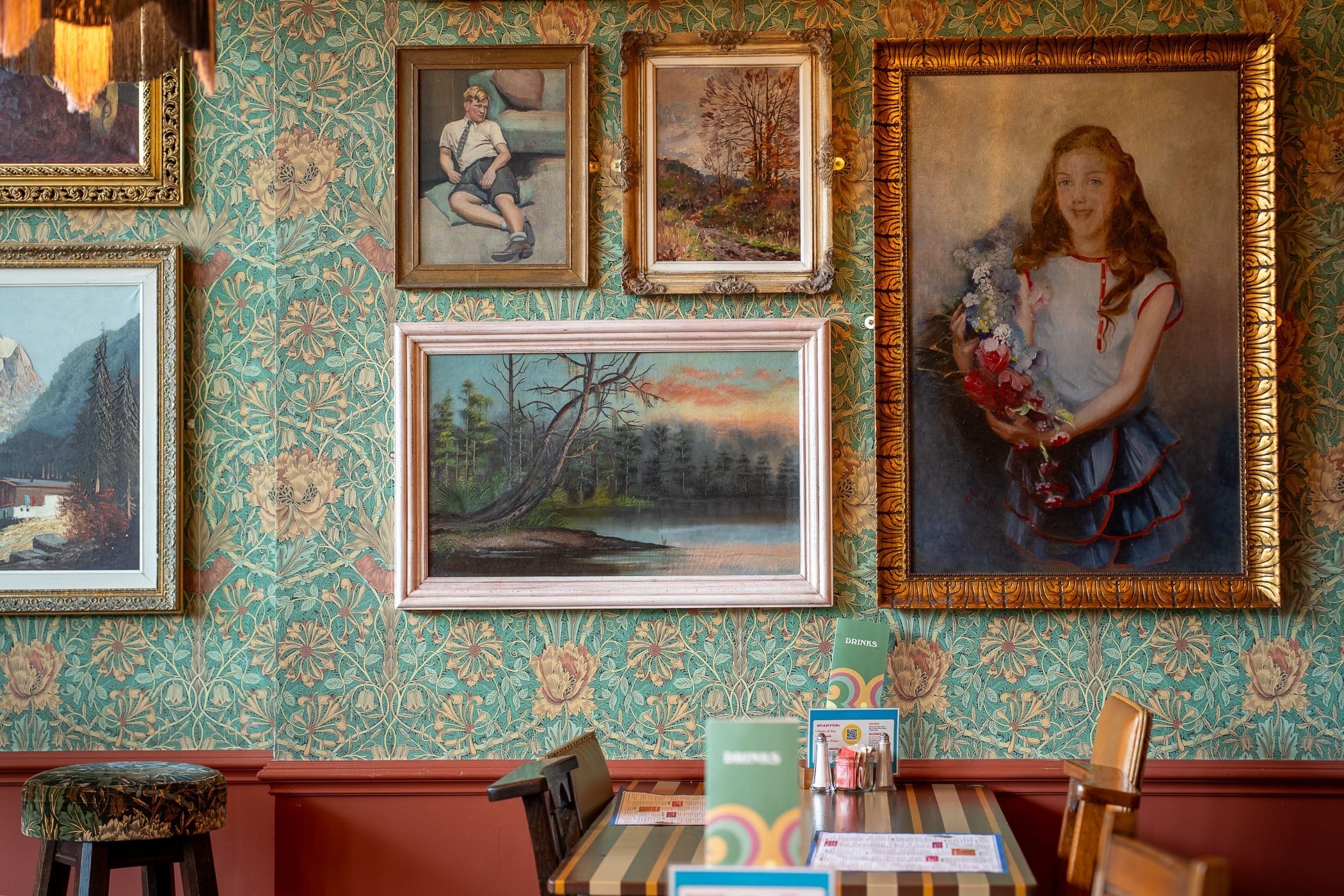 A cozy Pinto cafe with patterned green wallpaper, several framed paintings on the wall, and a table with menus and chairs in the foreground. The largest painting is a portrait of a smiling girl holding flowers.