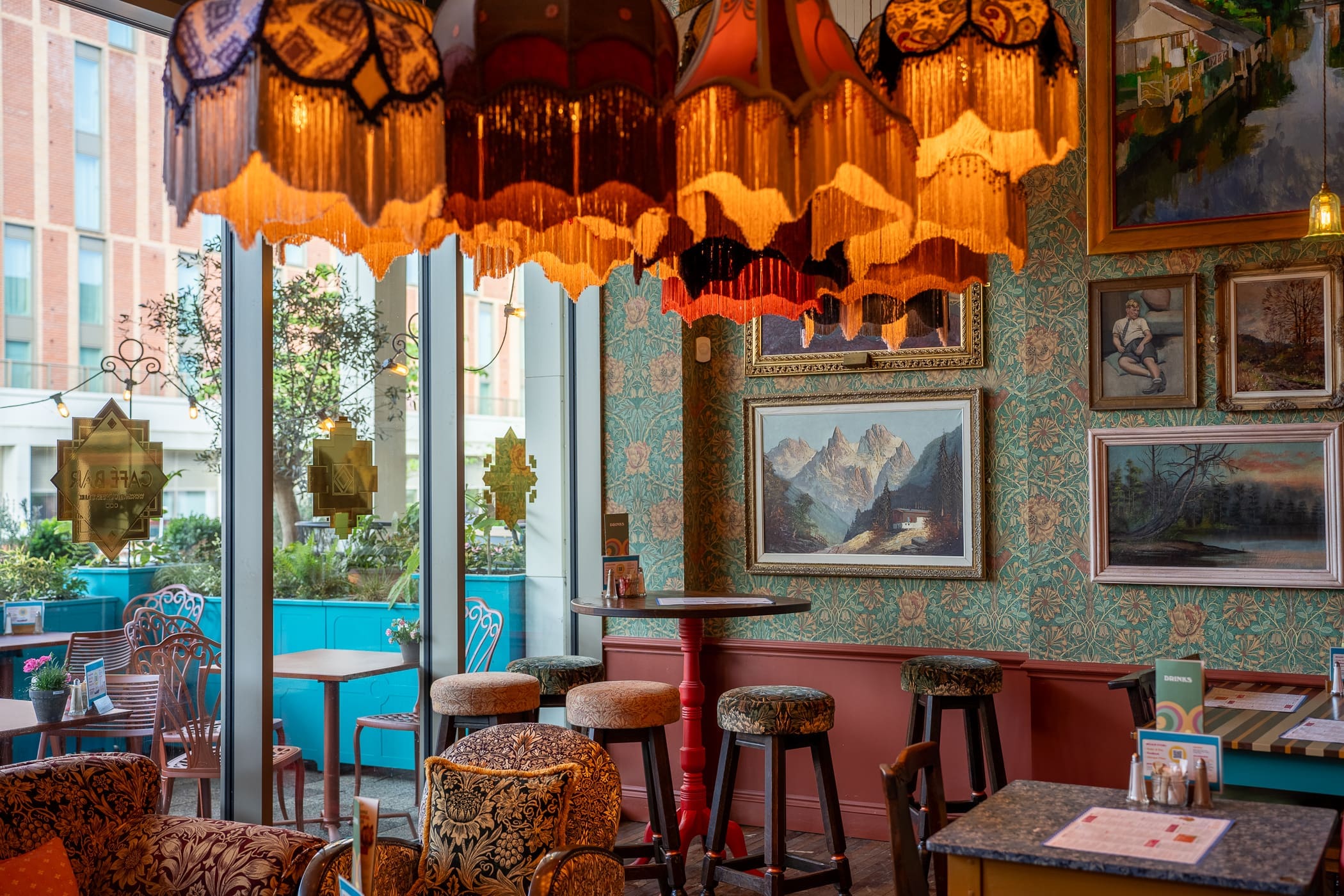Cozy vintage café interior with Pinto-patterned wallpaper, ornate lamps, paintings on the wall, cushioned stools, and large windows revealing outdoor seating and greenery. Warm lighting creates an inviting atmosphere.