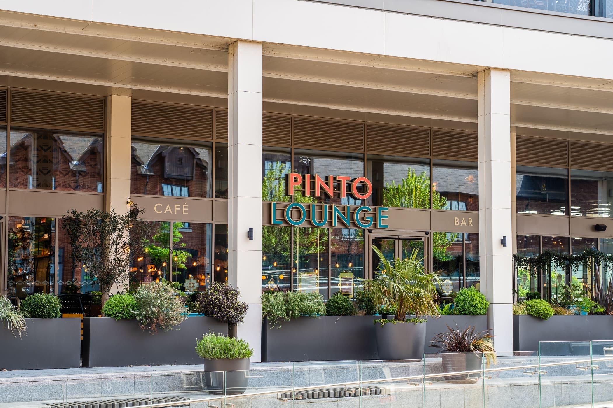 The exterior of Pinto Lounge café bar features large windows, lush potted plants, and modern Pinto signage. Reflections and rich greenery are visible through the glass, creating a welcoming atmosphere.
