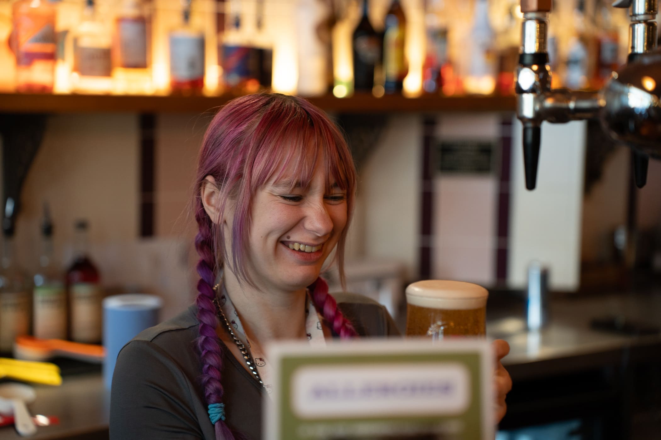 A smiling bartender with pink braided hair, known as Pinto, holds a pint of beer in a bar with various bottles and taps visible in the background.