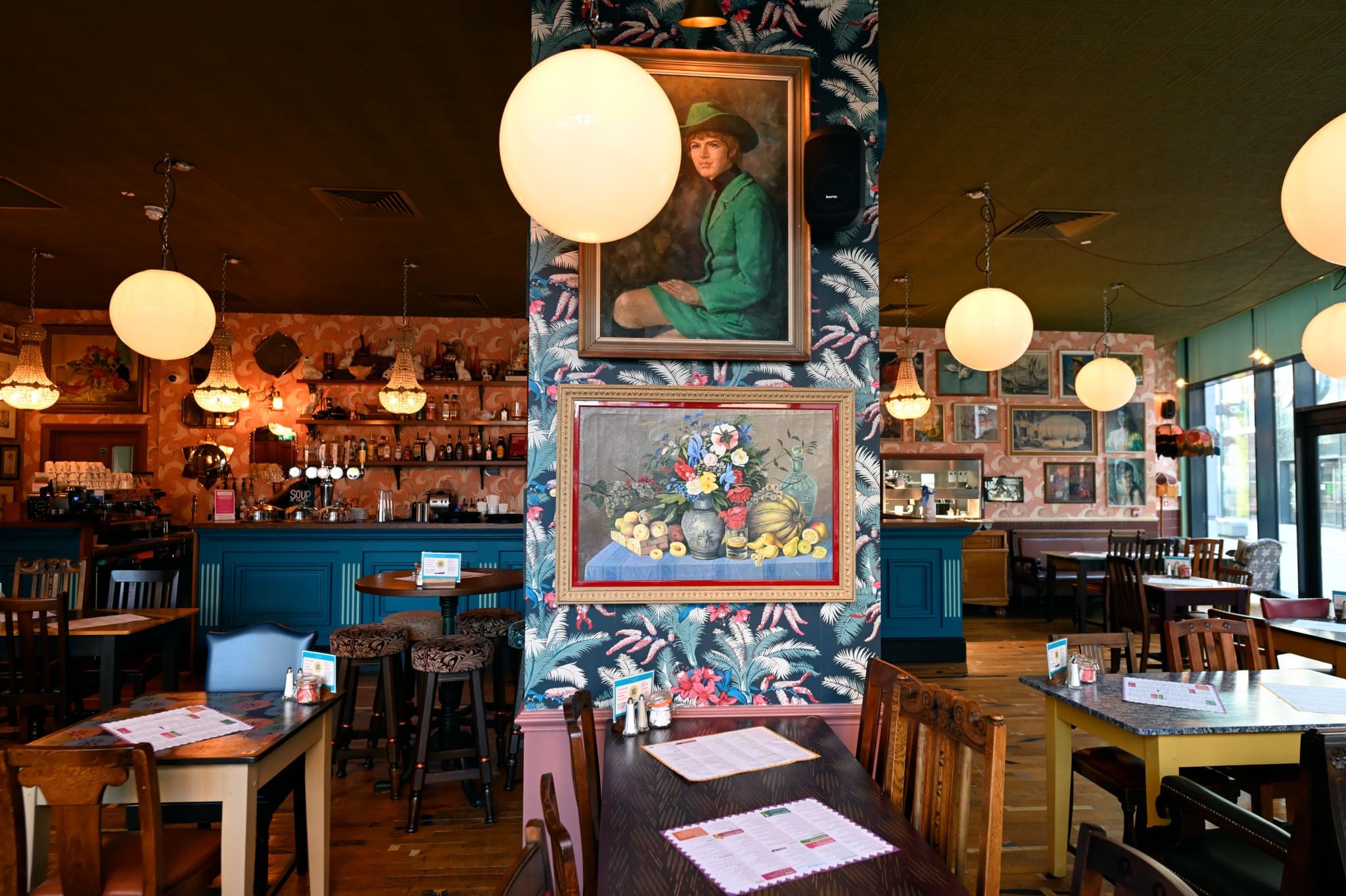 A cozy, colorful restaurant interior with eclectic vintage decor, round hanging lights, floral wallpaper, and paintings on the walls creates a pionero charm alongside wooden tables and chairs set for dining.
