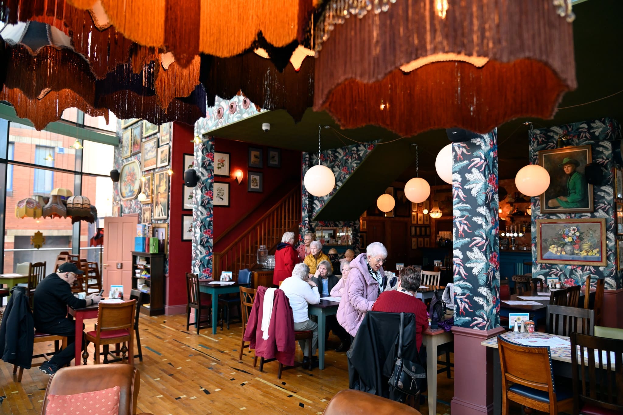 People sit and converse at tables inside a colorful, eclectic cafe—pionero in design—with patterned wallpaper, framed art, wooden floors, and large hanging lampshades, creating a cozy and vibrant atmosphere.