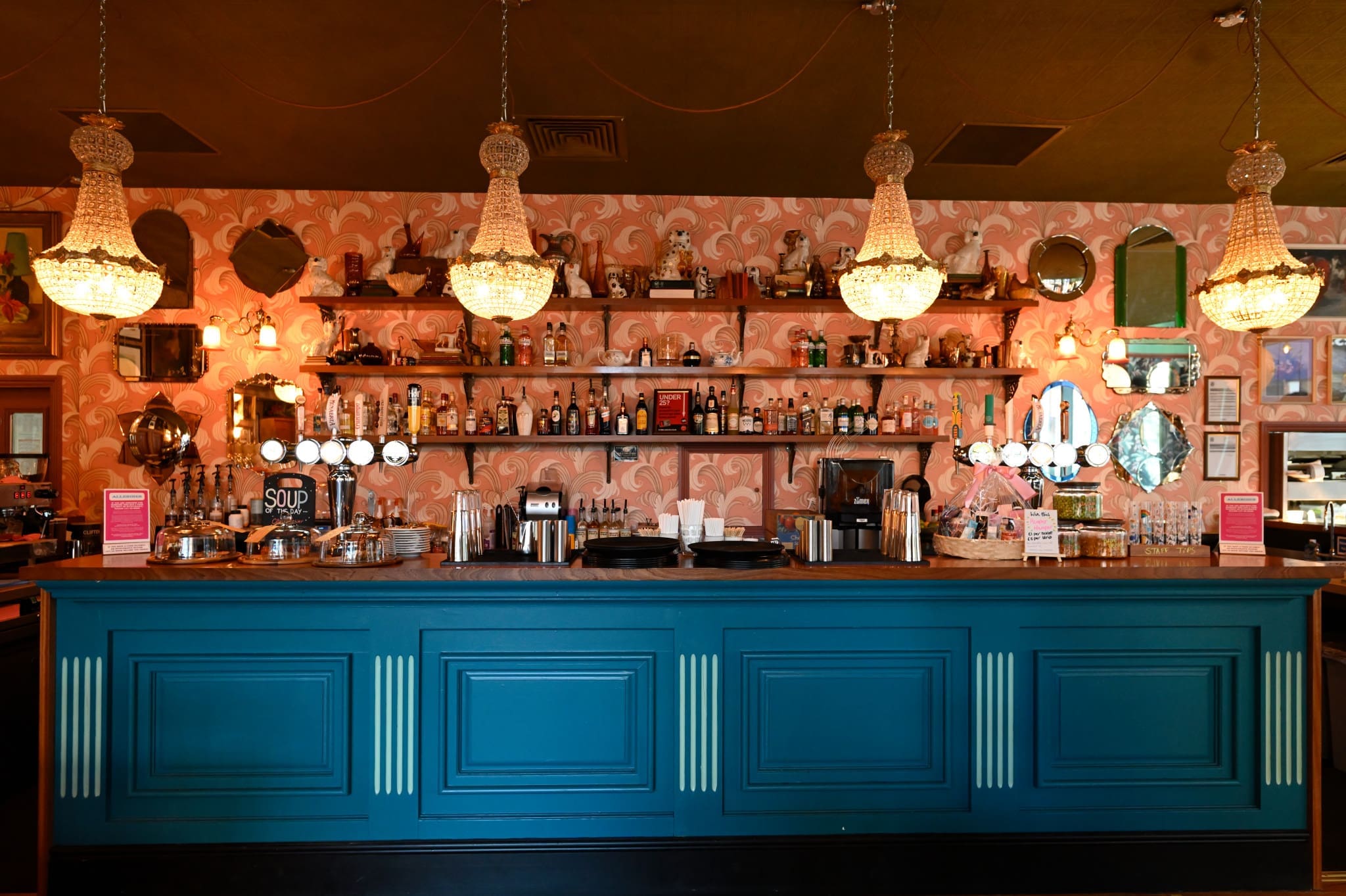 A stylish bar with a teal counter, ornate chandeliers, and patterned pink wallpaper. Shelves behind the bar display bottles and glasses, giving this pionero spot a cozy, vintage atmosphere with mirrors and lights enhancing the charm.