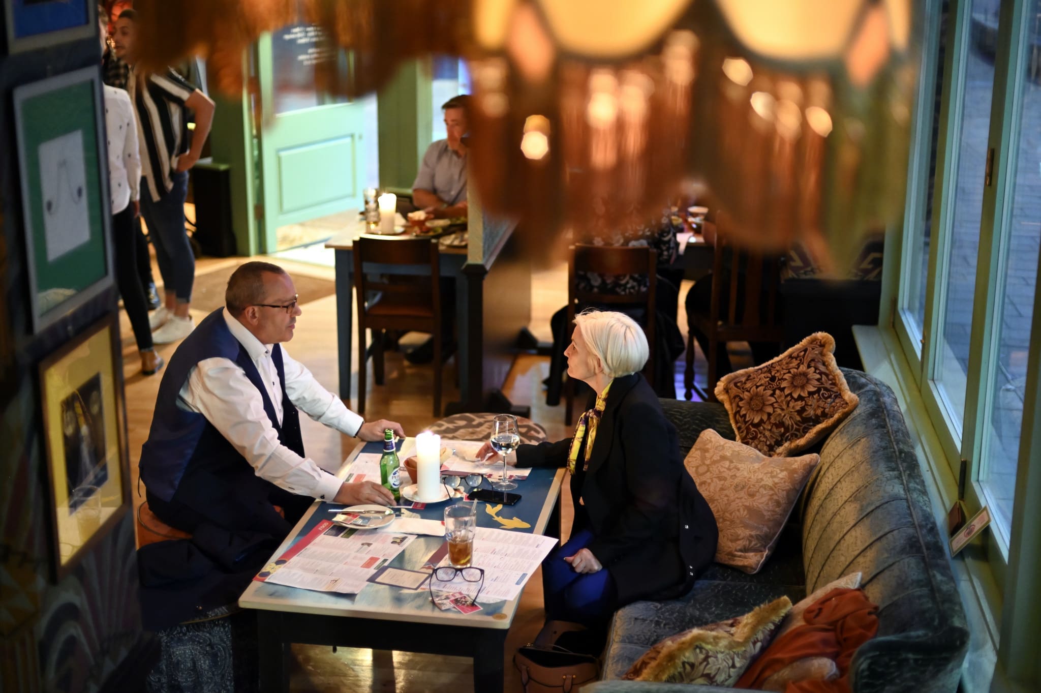A man and woman sit across from each other at a cozy Ponto restaurant table with drinks, papers, and a tablet. Warm lighting and plush seating create an intimate atmosphere, while other diners can be seen in the background.