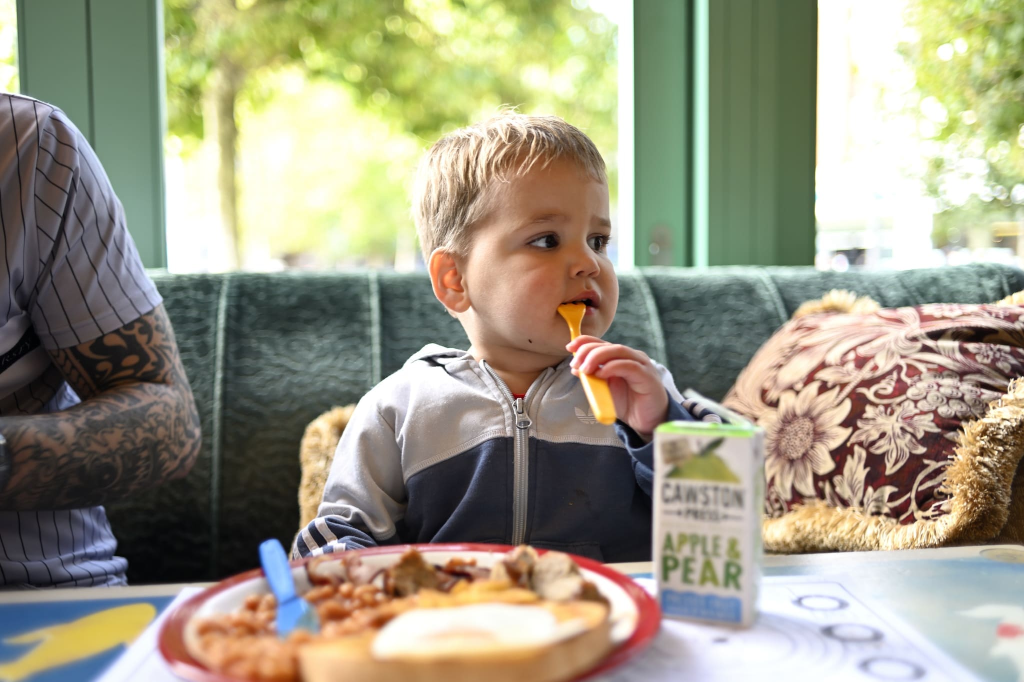 A young child sits at a table eating with a yellow fork, next to a plate of food and a Ponto apple & pear juice box. The child wears a jacket and looks to the side, with sunlight coming through windows behind them.