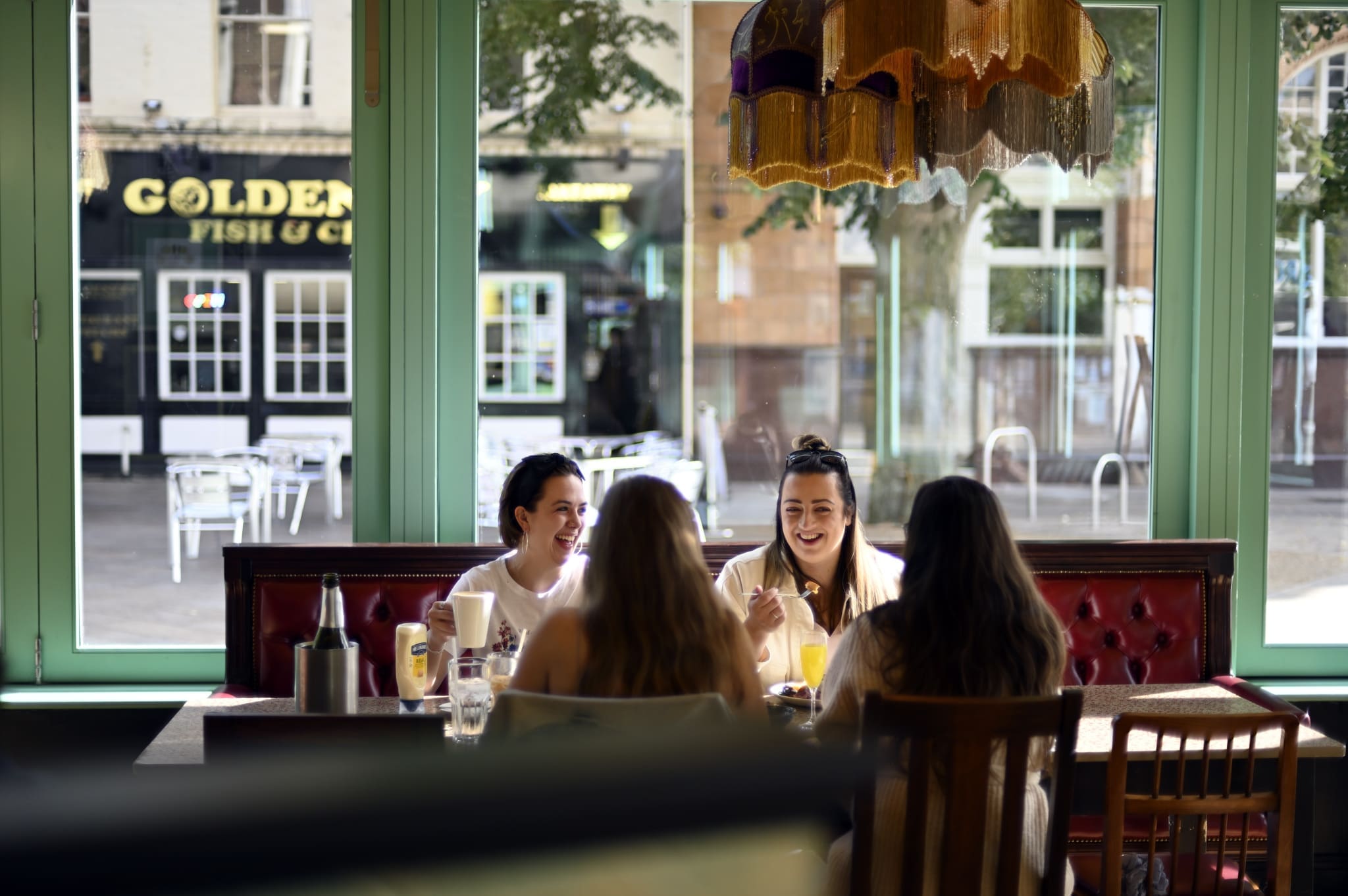 Four women sit together at a table inside the cozy Ponto cafe, talking and smiling. Large windows behind them reveal a street view with trees and the "Golden Fish & Chips" shop across the road.
