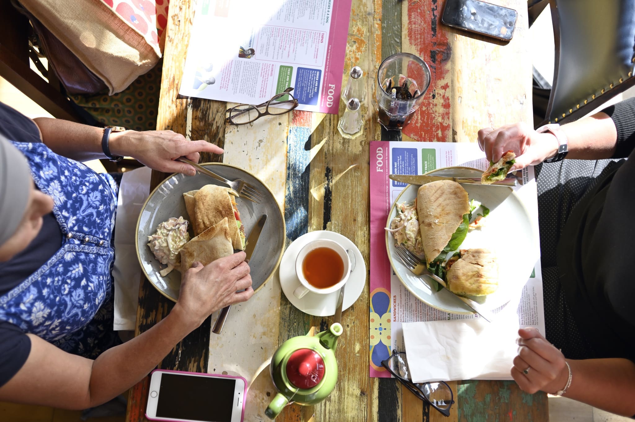 Two people sit at a colorful wooden table at Ponto, enjoying sandwiches with coleslaw, tea, and soda. Menus, glasses, and a phone are scattered across the table. The scene is bright and casual, captured from above.