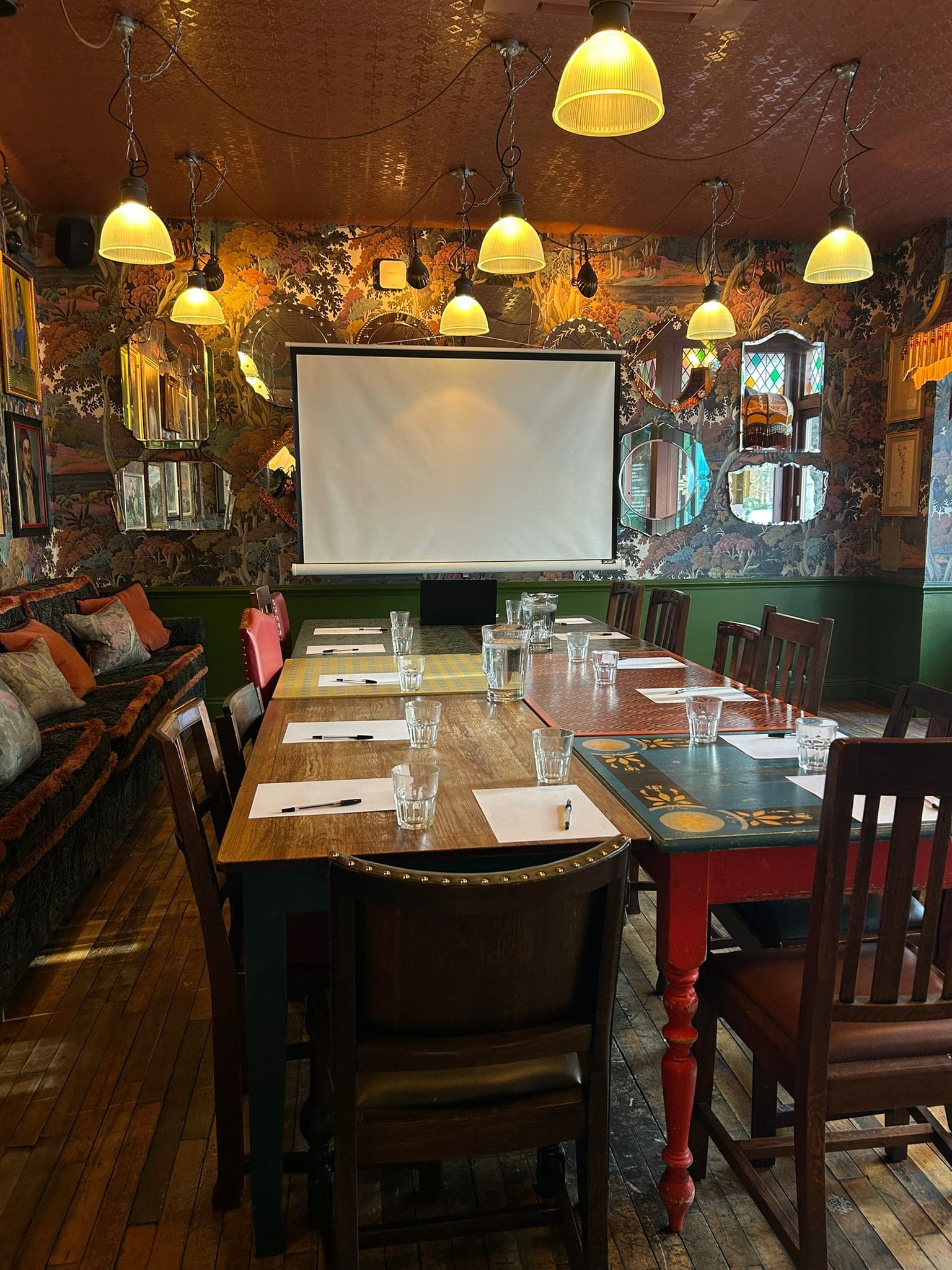 A cozy room with a long wooden table set for a meeting, featuring notepads, glasses, and pens. There's a projector screen at the end, warm lighting, patterned wallpaper, and eclectic decor on the walls.