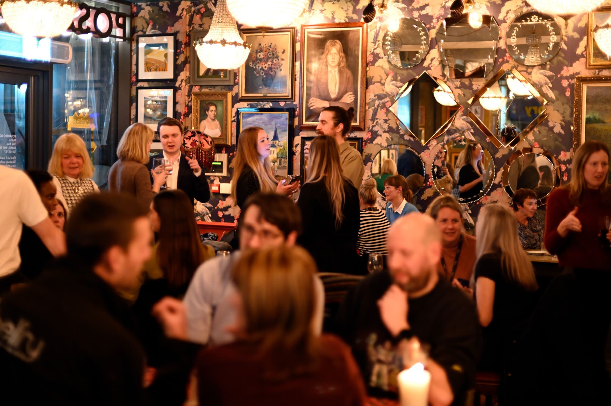 A lively group of people socialize and dine in a warmly lit Posadero restaurant adorned with ornate mirrors, framed art, and floral wallpaper. Some guests are standing and chatting while others sit at tables.