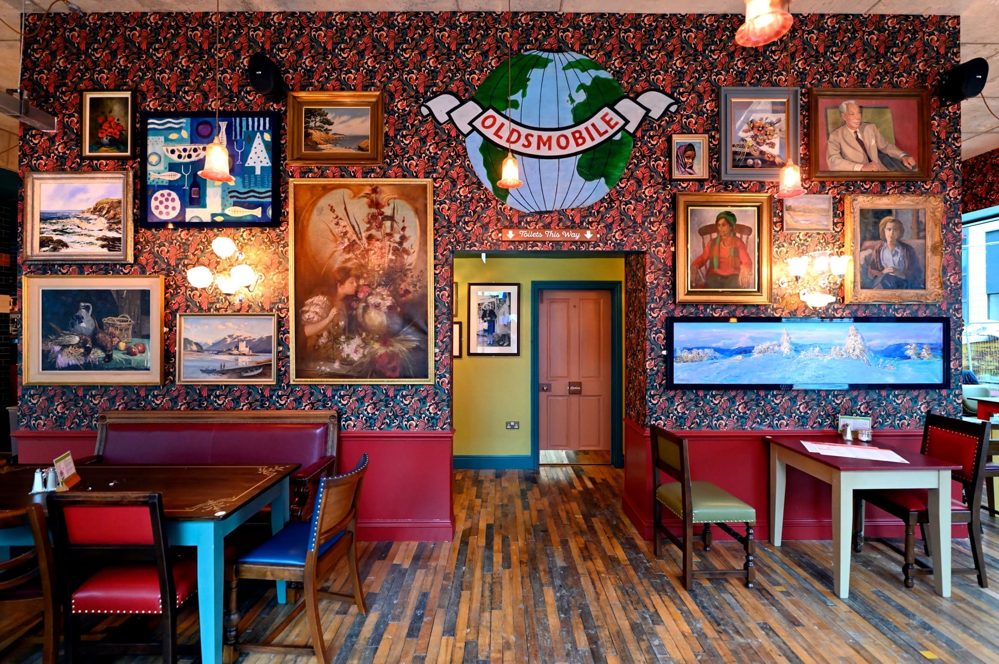 Eclectic Posadero restaurant interior with colorful mismatched chairs, wooden tables, and a wall decorated with vintage paintings, framed art, and an “Oldsmobile” globe sign on floral wallpaper. Warm lighting creates a cozy ambiance.