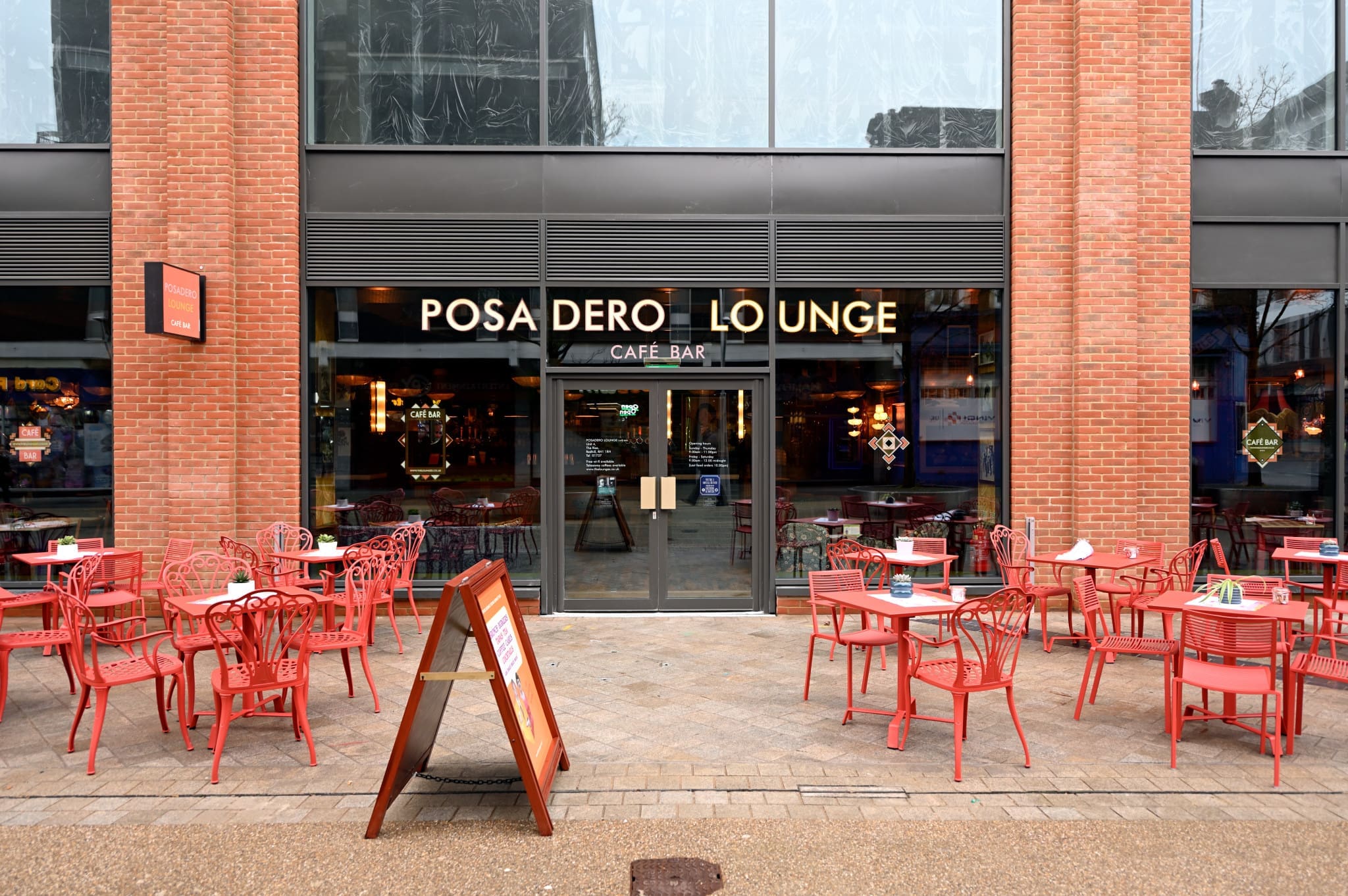 Outdoor seating area with red chairs and tables in front of Posadero Lounge, a café bar set in a modern brick building with large windows and glass doors.