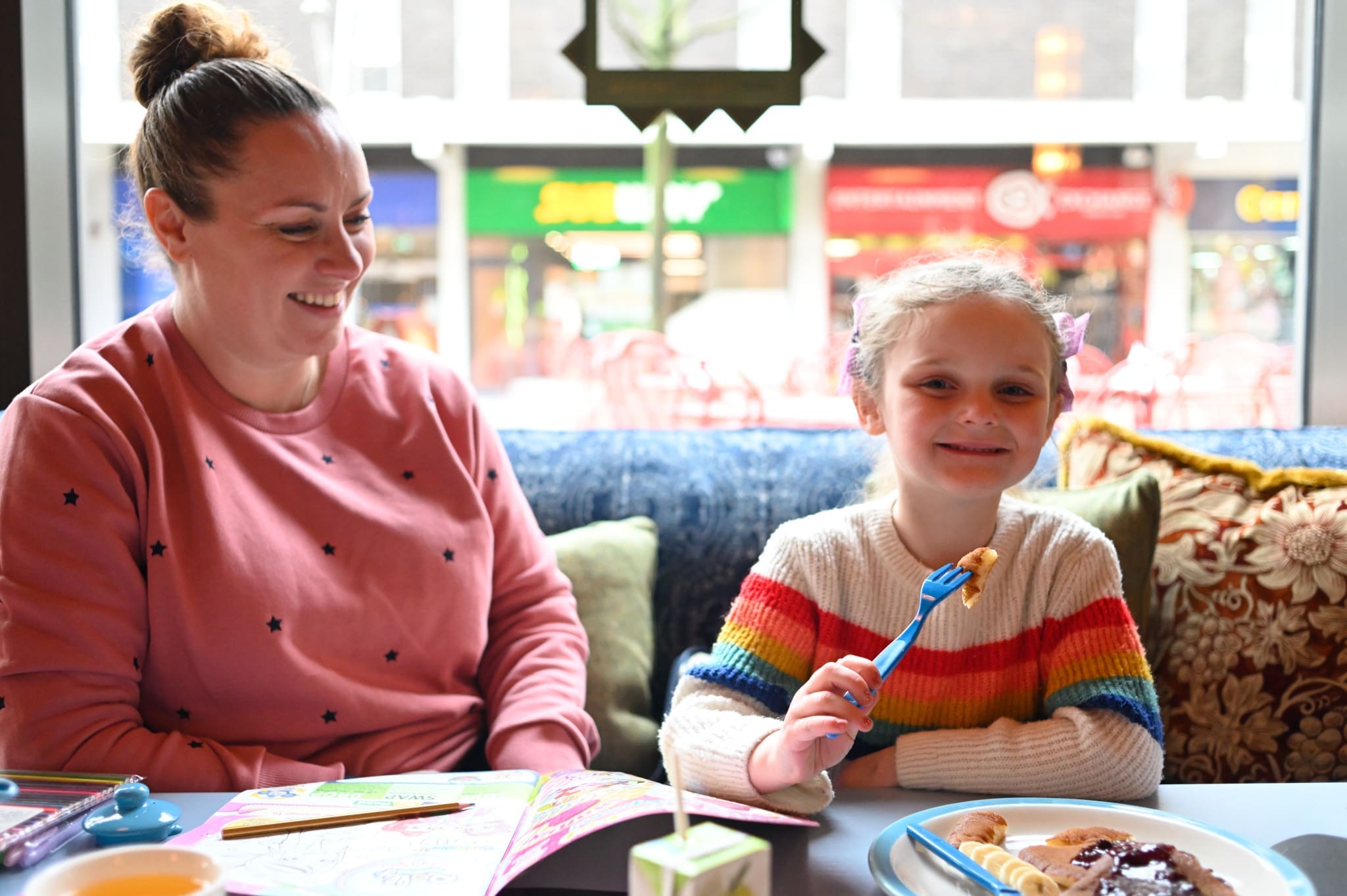 A woman and a smiling young girl sit at a table in Posadero. The girl holds a fork with food, and plates and drinks are in front of them. The woman looks at the girl, both appearing happy and relaxed.