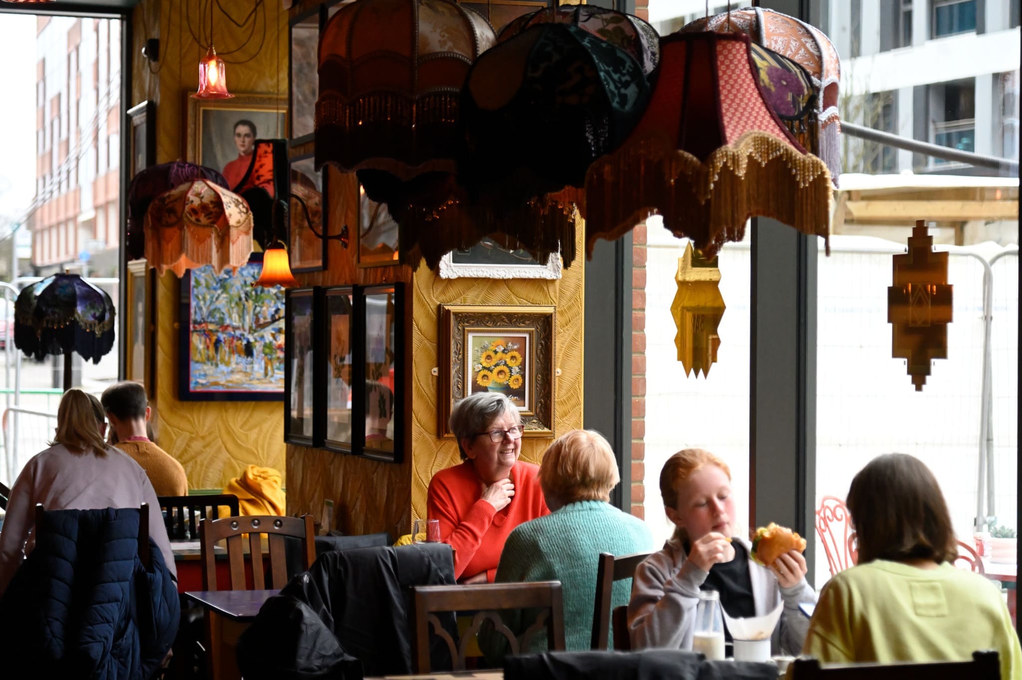 People sit and eat at tables inside a cozy, warmly lit café called Posadero, decorated with colorful lamps, paintings, and large windows that reveal an urban street outside.