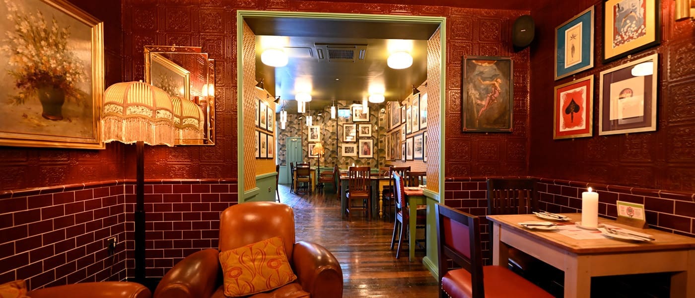 A cozy, warmly lit Postino restaurant with vintage decor, leather chairs, dark wood floors, and walls adorned with framed art and photographs. The dining area is visible through an open doorway.