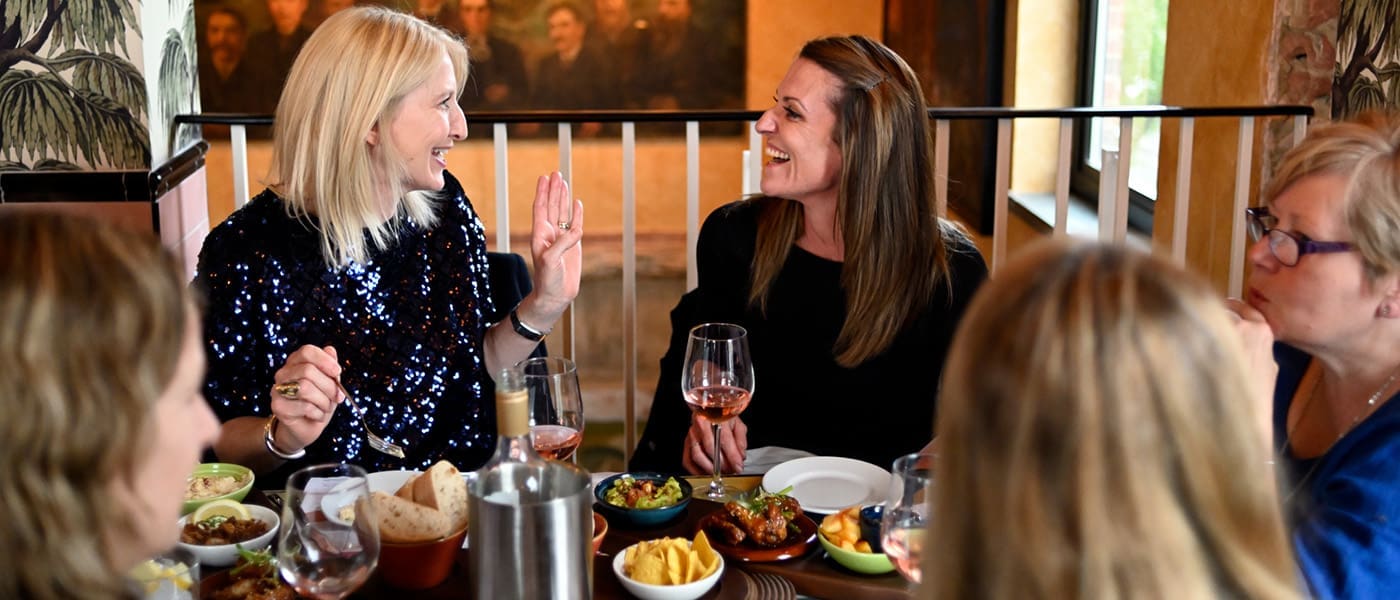 Five women sit around a lively table at Postino, enjoying delicious food and drinks. Two women in the center are smiling and chatting, with various dishes and glasses of wine creating a warm, inviting atmosphere.