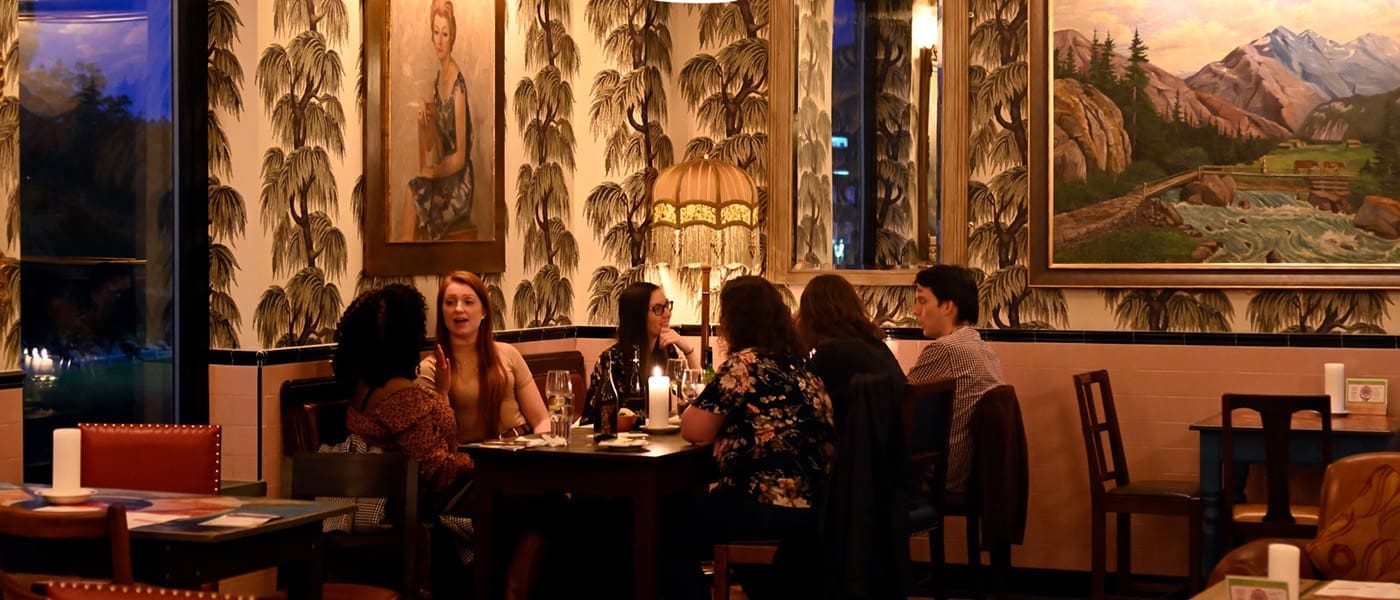 A group of six people sit at a table in Postino, a stylish, warmly lit restaurant with vintage decor, wallpaper, and large landscape paintings on the walls, engaged in conversation and dining together.